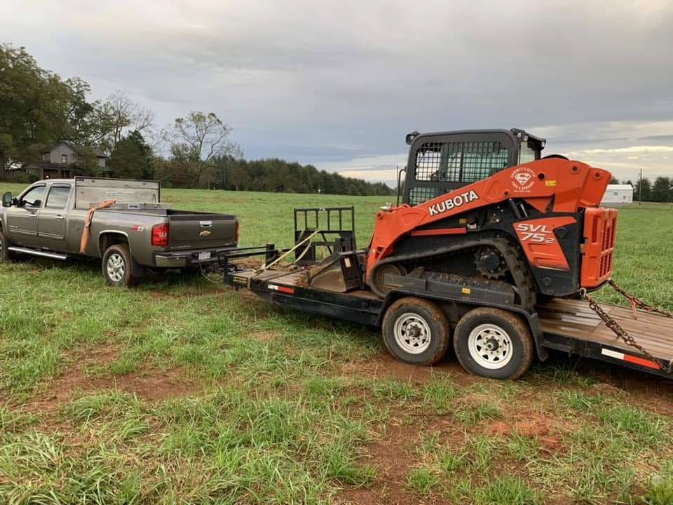 A gray pickup truck towing a flatbed trailer with an orange Kubota skid steer on a grassy field.