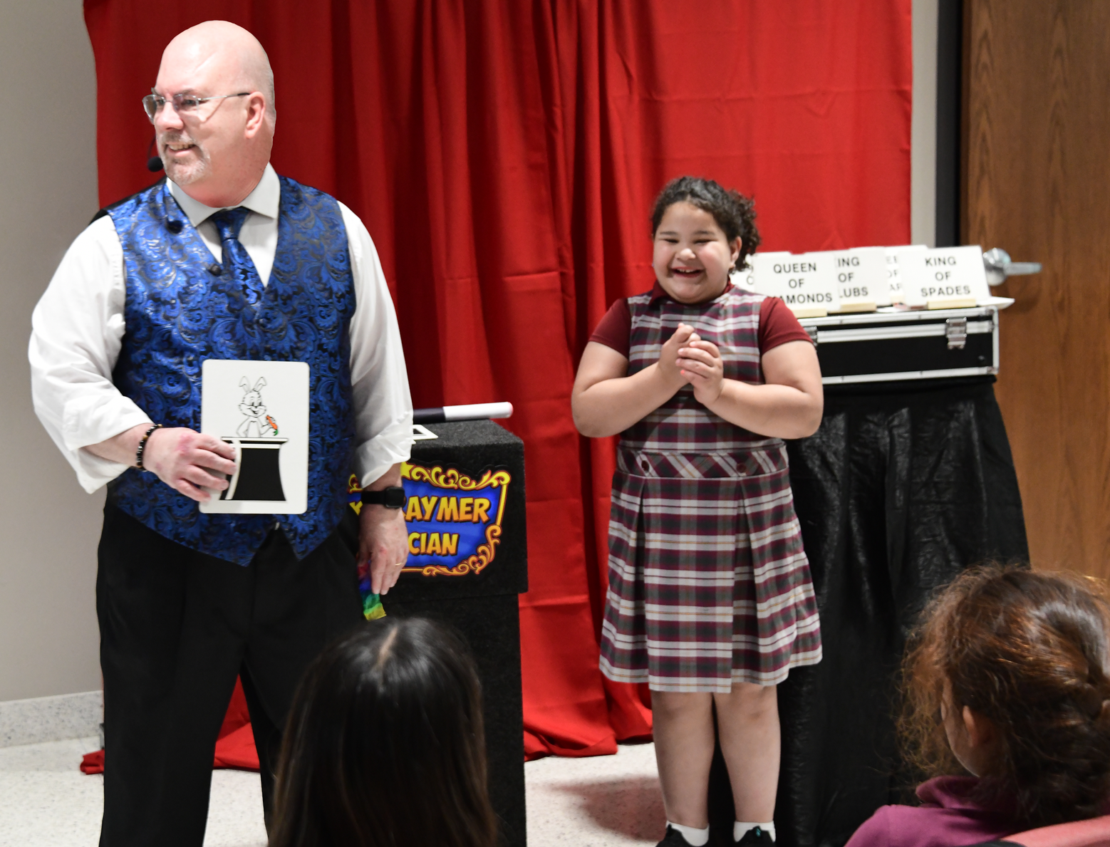 Magician with blue vest holds a card; girl in plaid dress smiles, watching the magic show.