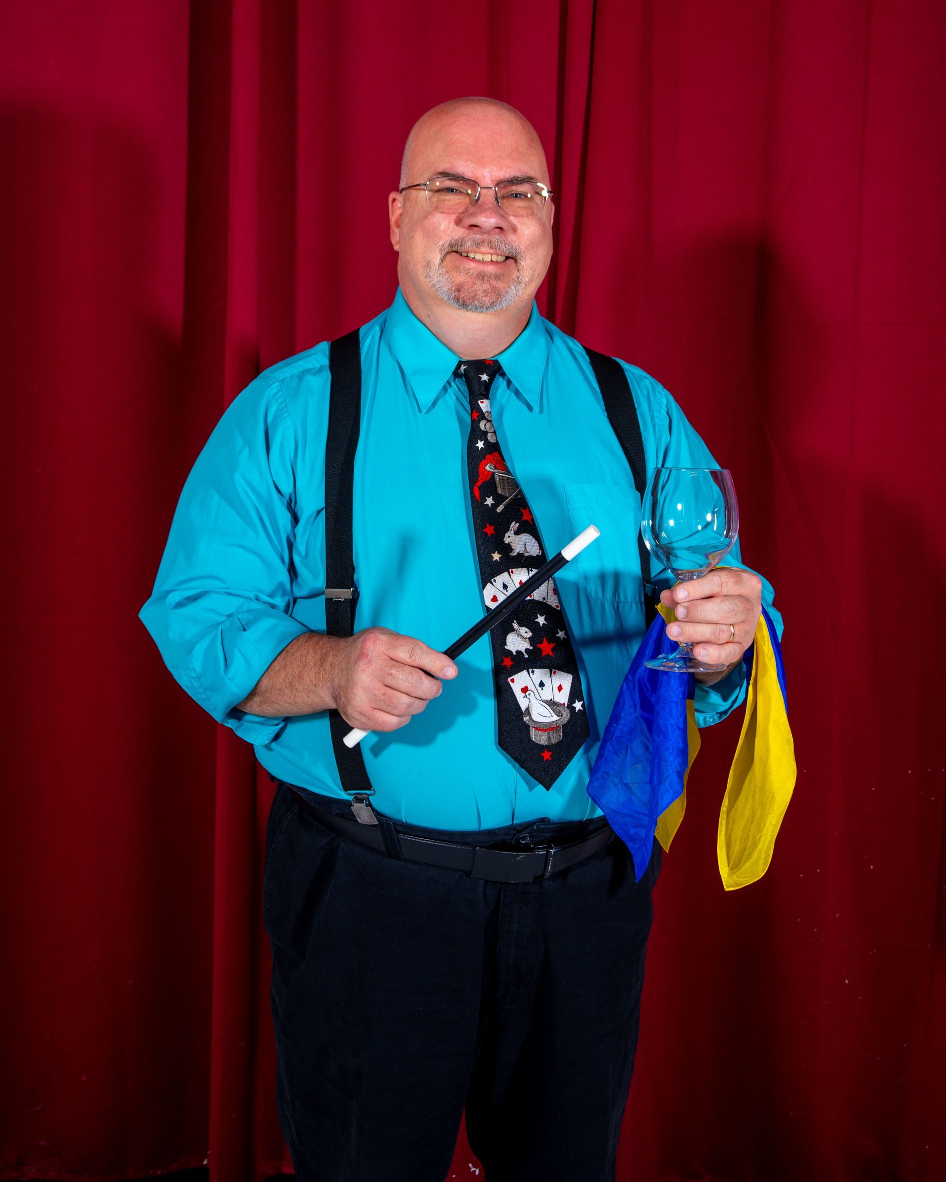 man in teal shirt and black pants holds a magic wand a glass and a blue and yellow colored handkerchief