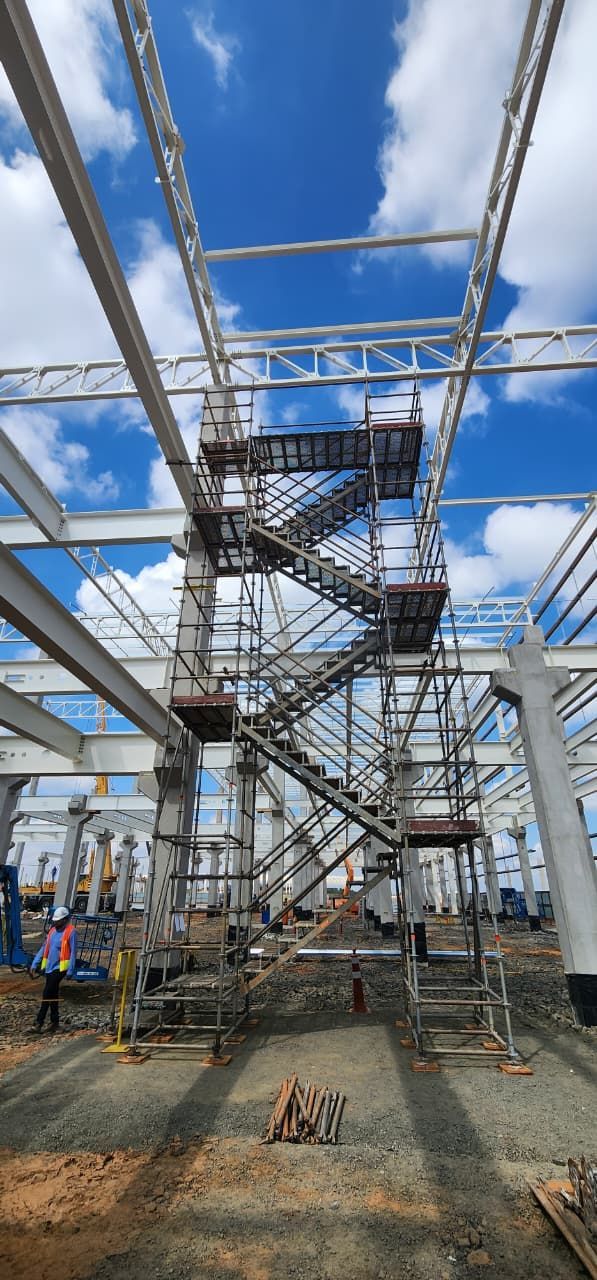 Canteiro de obras com estrutura metálica, andaimes e escada contra um céu azul com nuvens.