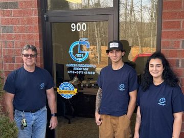 A person wearing overalls works on plumbing in a bathroom. They stand on a toilet with a toolbox in front.