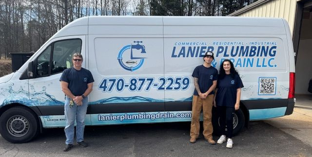 A person wearing overalls works on plumbing in a bathroom. They stand on a toilet with a toolbox in front.