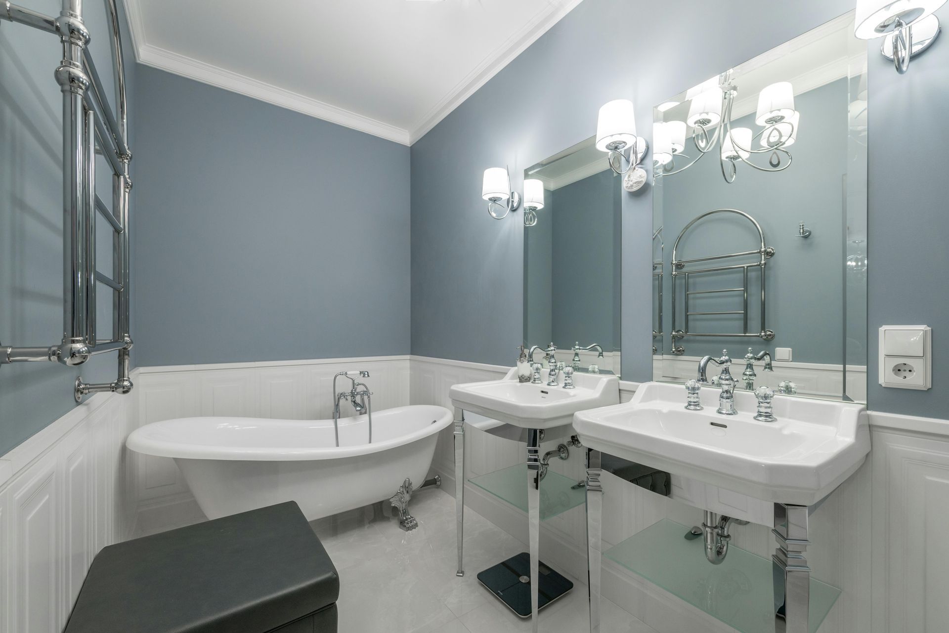 Elegant bathroom with a blue wall and white trim. Features a clawfoot tub, pedestal sinks, and a large mirror with decorative lighting.