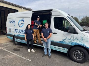 Three staff members stand beside a white plumbing van with blue branding, parked outside an industrial building.