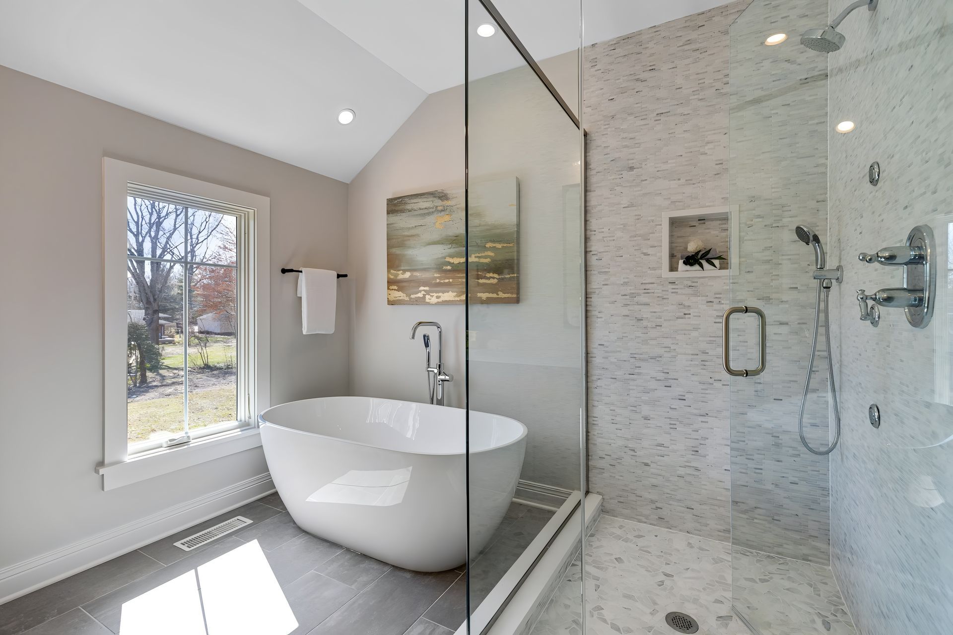 A modern bathroom featuring a white, freestanding tub, a glass-enclosed shower, and a window overlooking a yard.