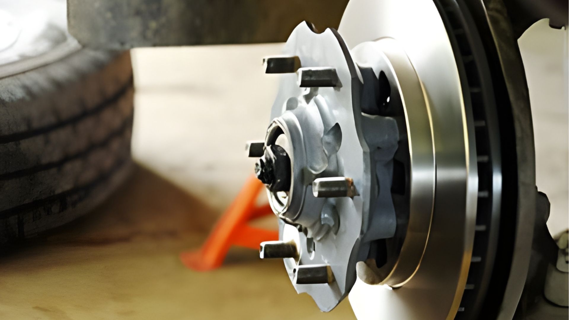 A close-up of an automotive wheel hub and brake rotor installed on a vehicle, with a tire and jack stand in the background.