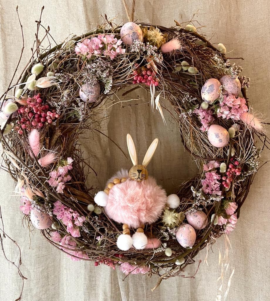 Une couronne avec des fleurs roses et des œufs et un lapin en peluche au milieu