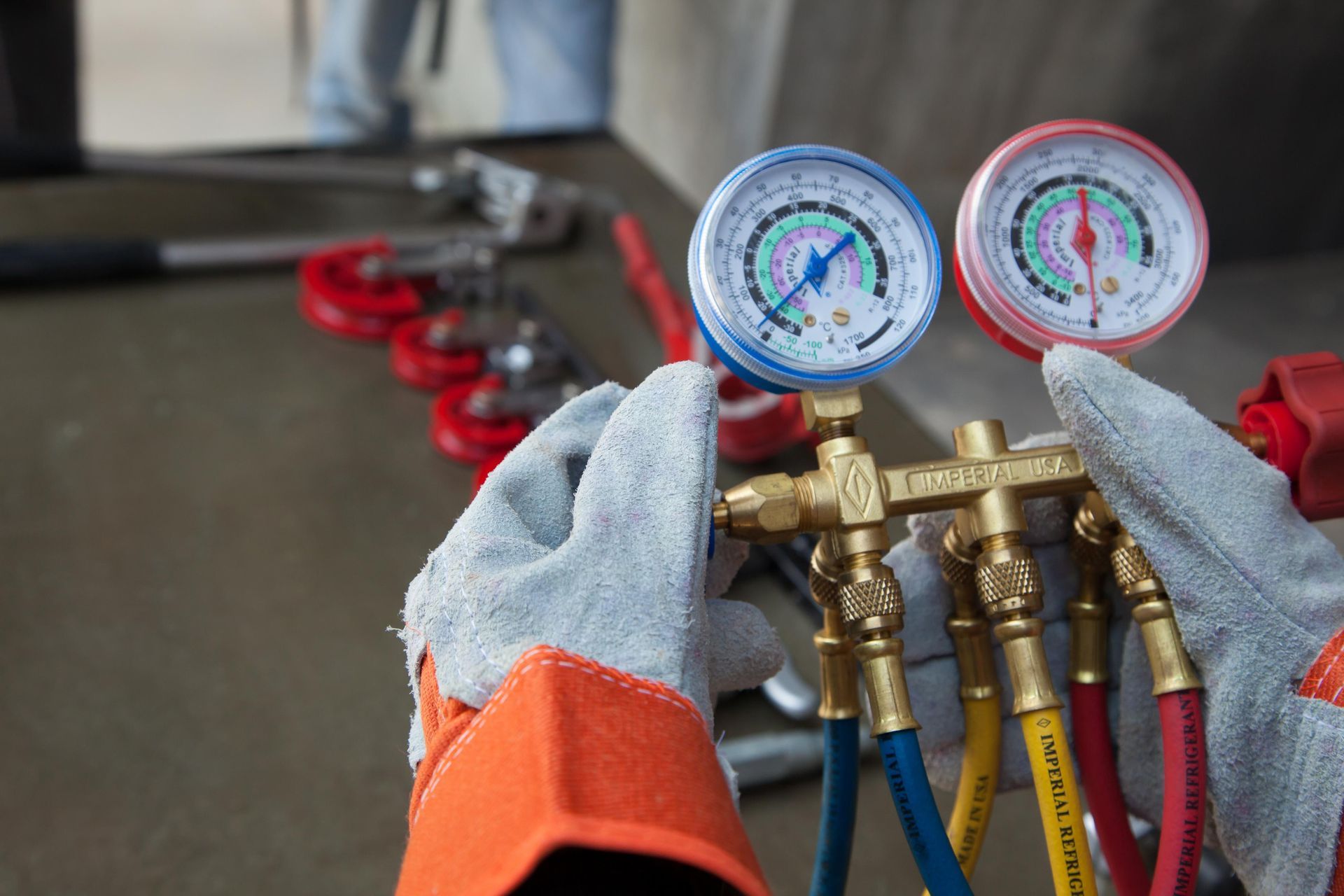 Hands in gloves holding HVAC manifold gauges with tubing; tools in background.