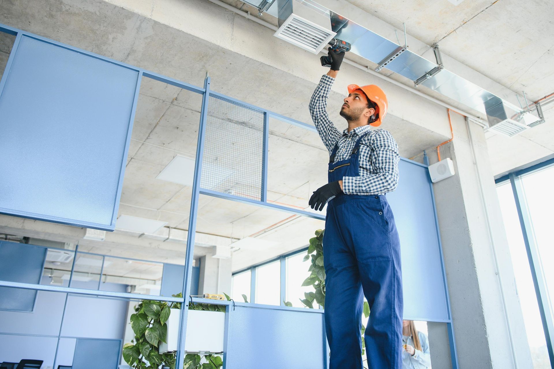 Construction worker in blue overalls and orange hard hat working on ceiling ventilation ducts.