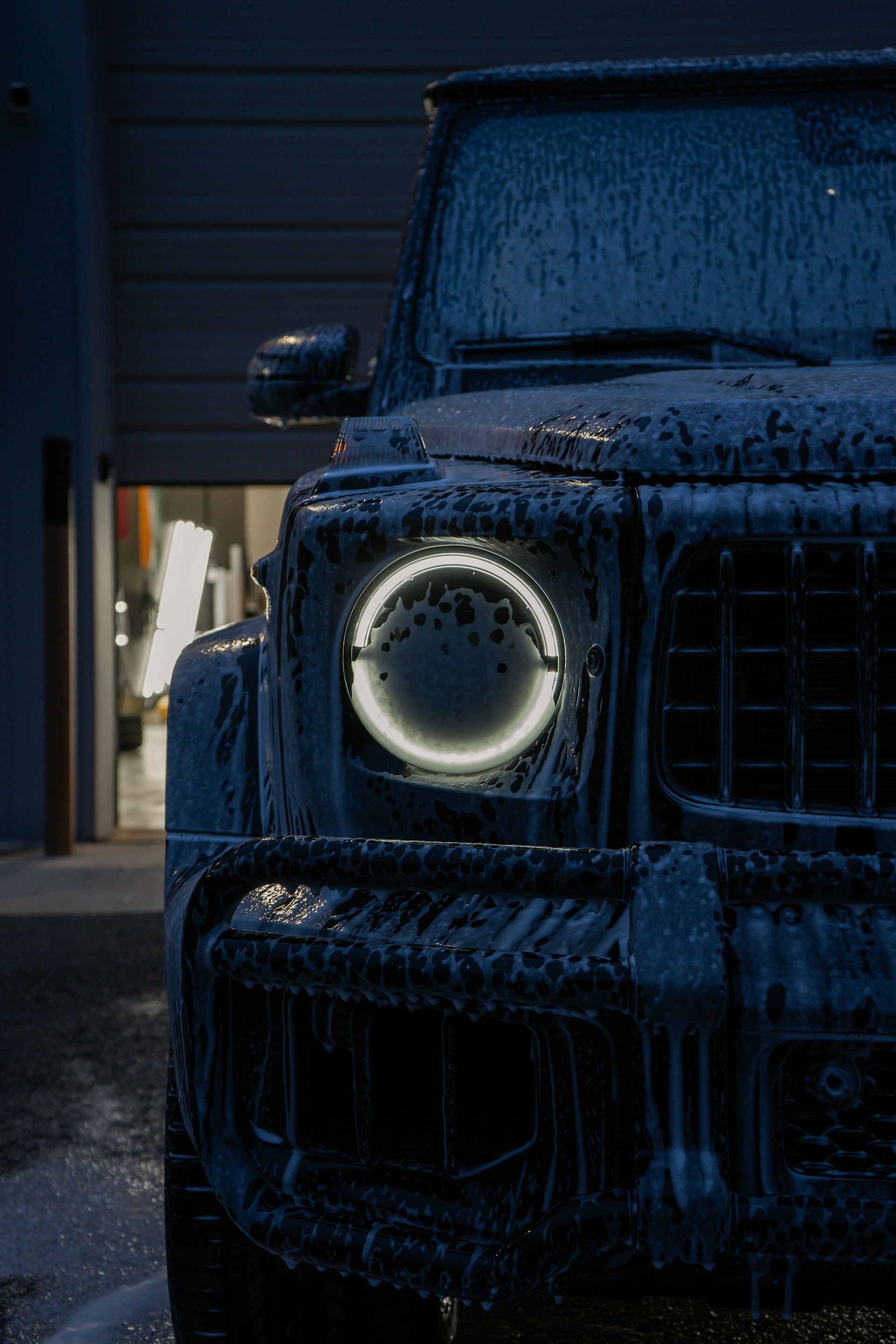 A Mercedes-Benz G-Wagon covered in thick, dark blue car wash soap with its circular headlight glowing in a garage.