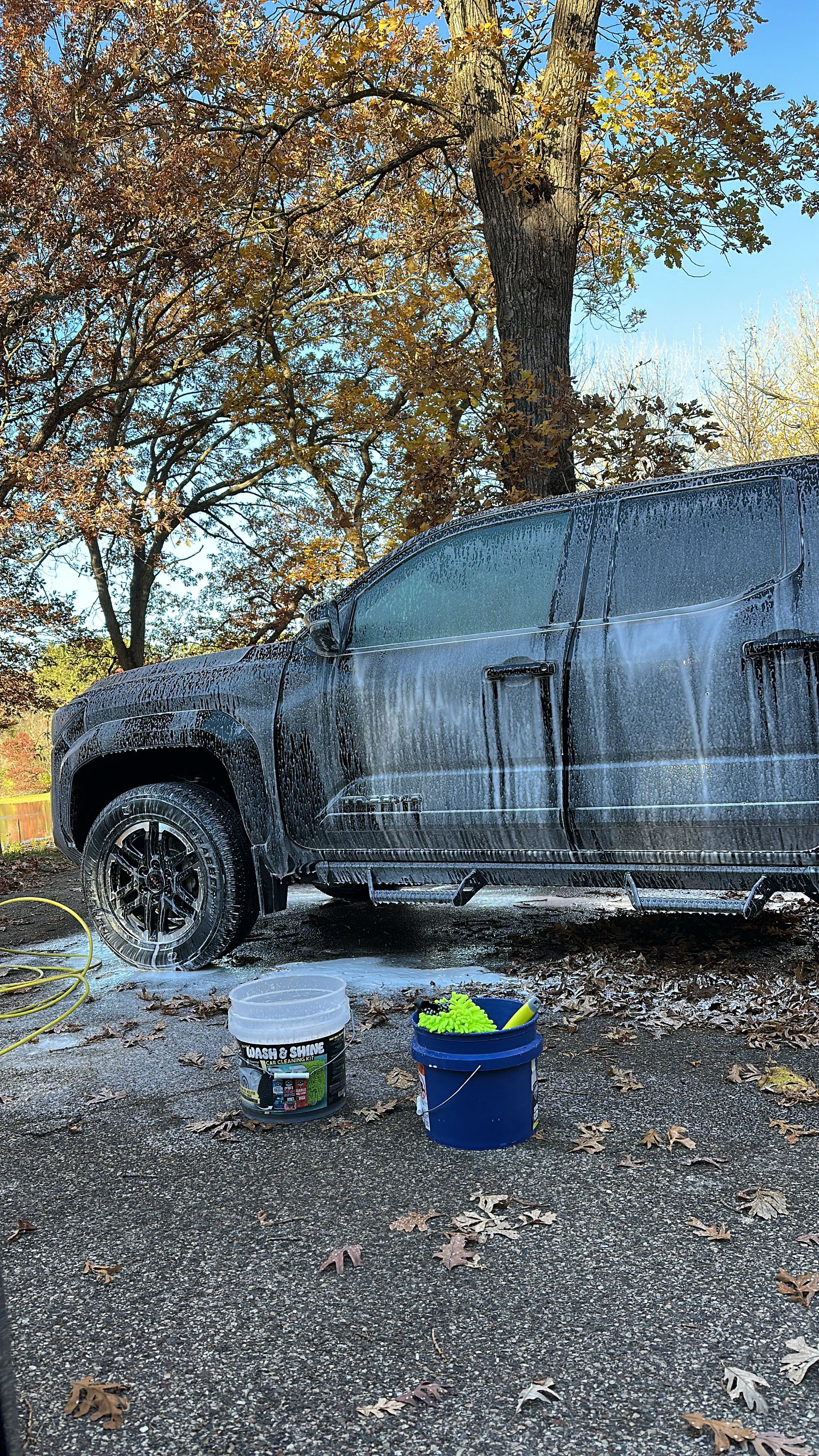 A dark pickup truck covered in white soap suds parked on a gravel surface outdoors under autumn trees.