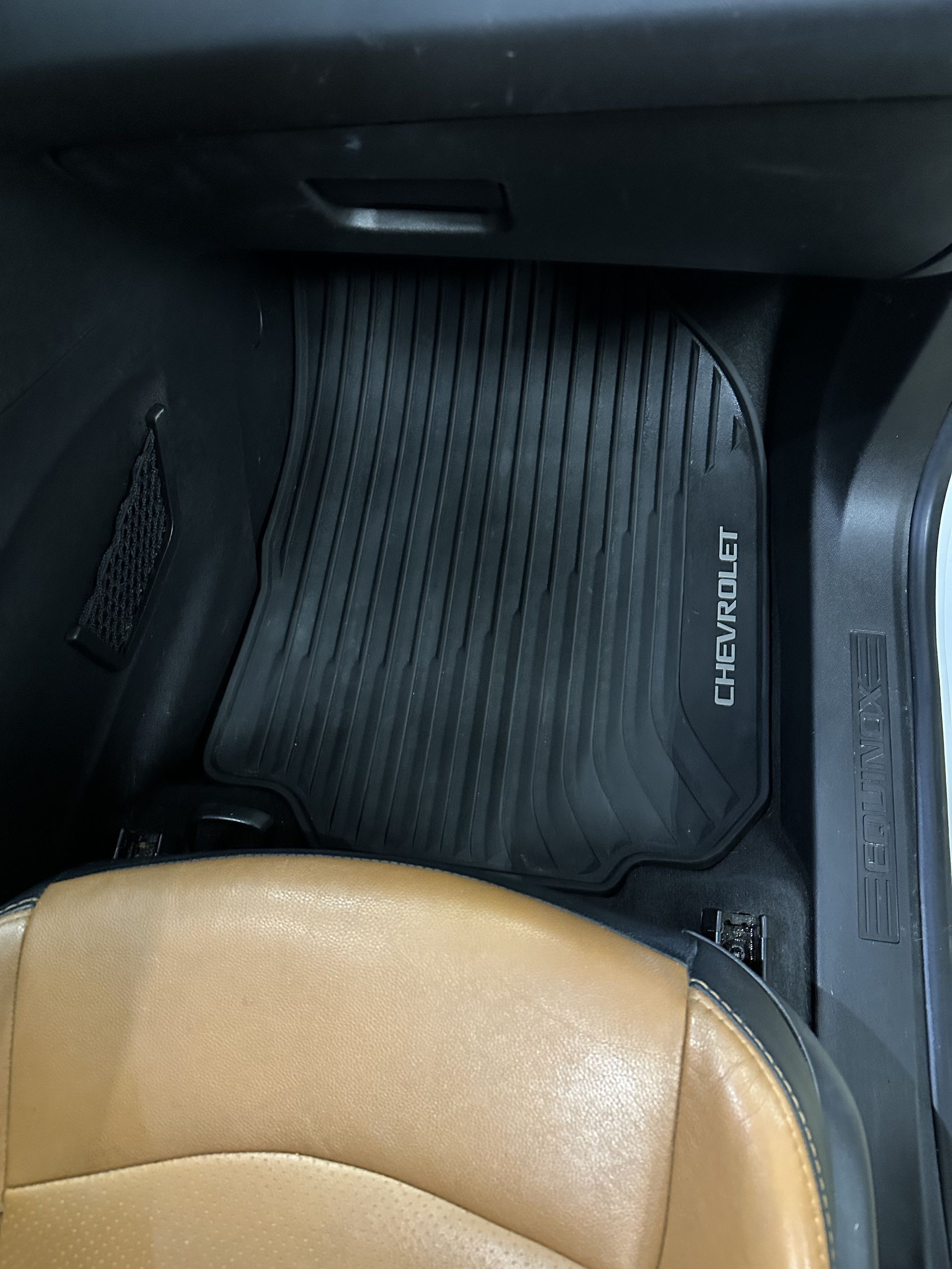 A black, ridged rubber Chevrolet floor mat sits on the floorboard of a car interior with tan seating.