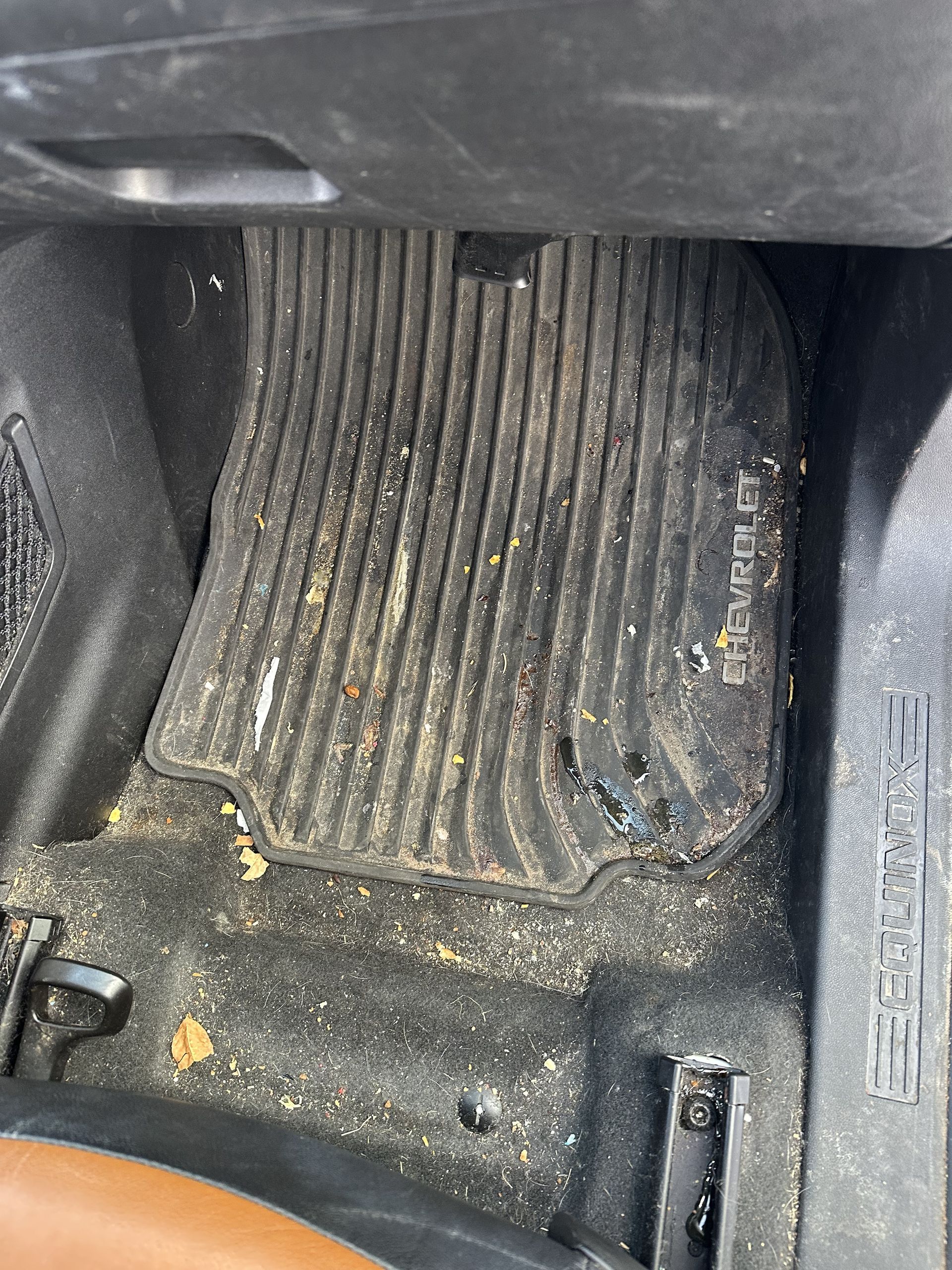 Dirty black rubber floor mat inside a vehicle footwell, surrounded by loose debris and dirt on the carpeted floor.