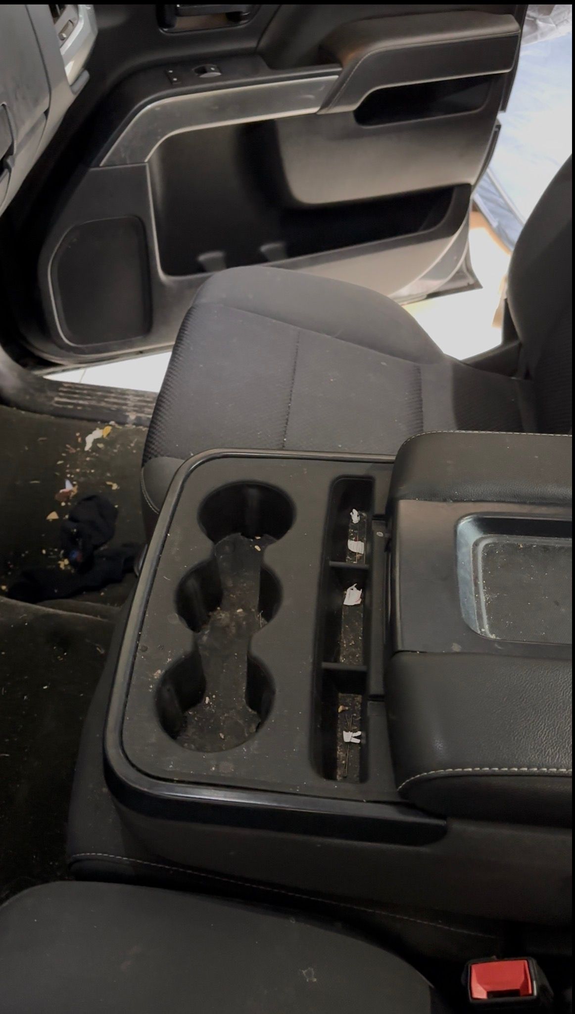 The interior of a vehicle shows a dark center console with three cup holders and a storage area covered in debris.