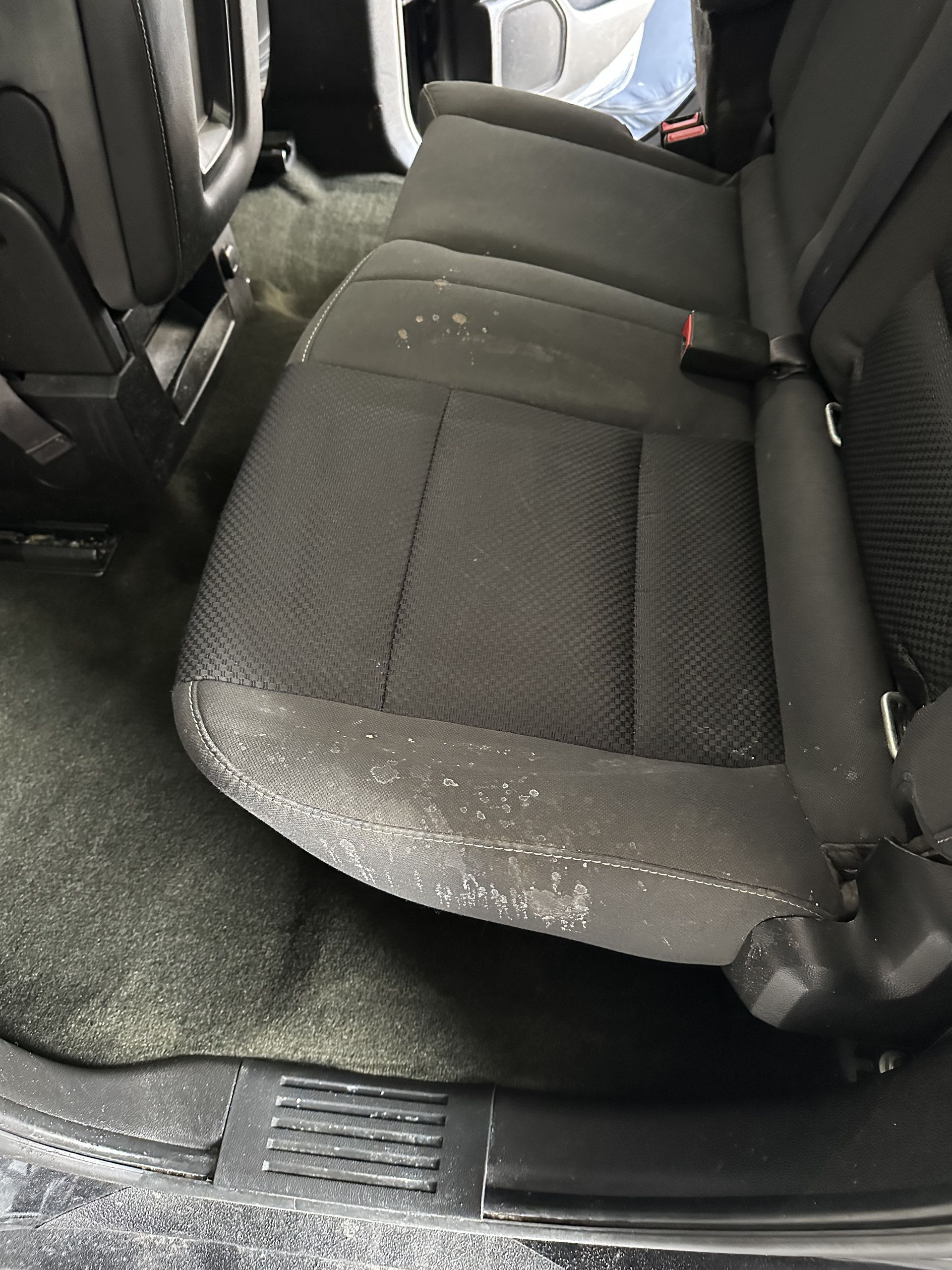 A car's black fabric backseat featuring light-colored stains, crumbs, and debris scattered across the seat cushion.