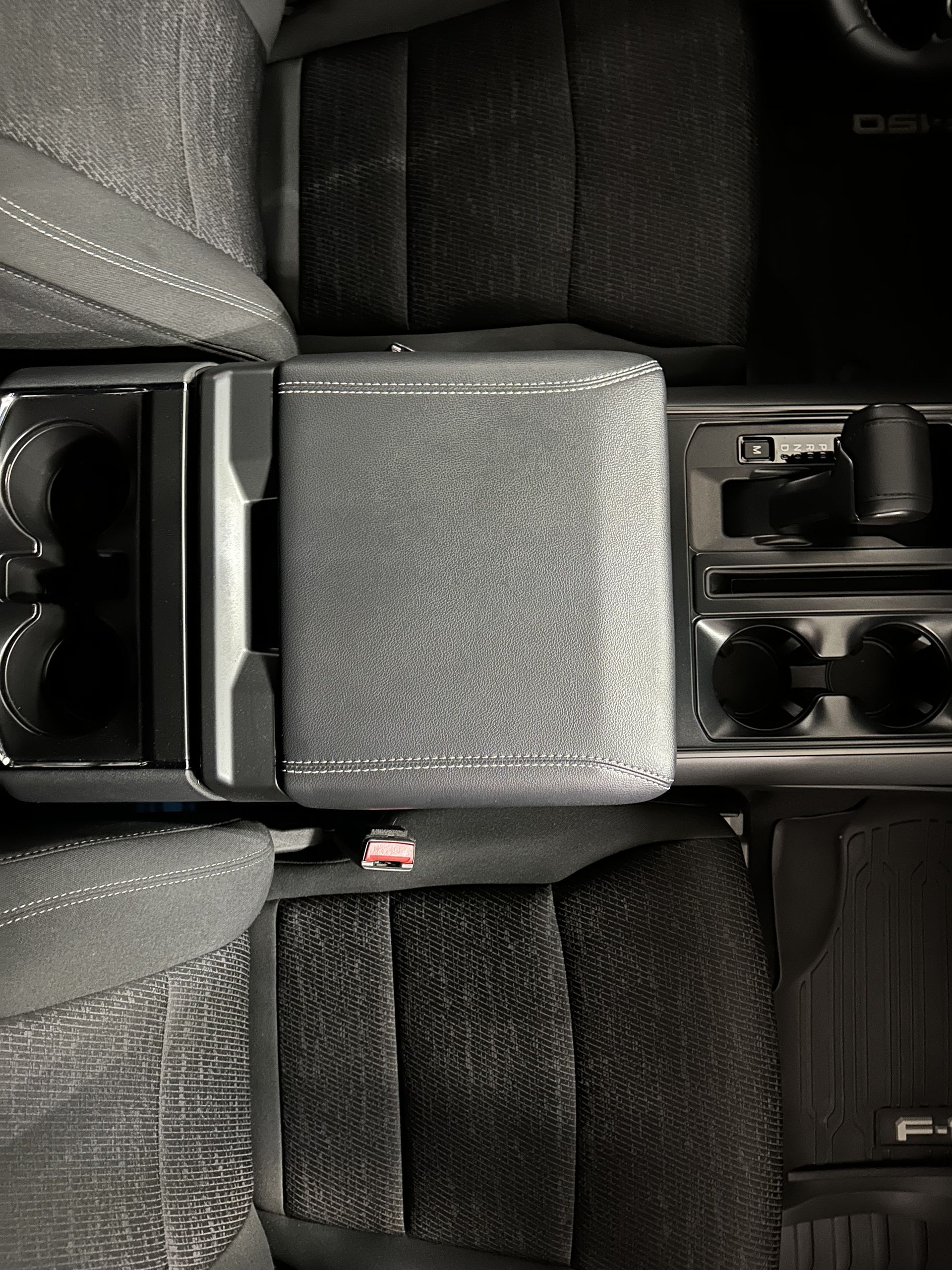 Top-down view of a car center console featuring a textured storage lid, cup holders, and portions of gray fabric seats.