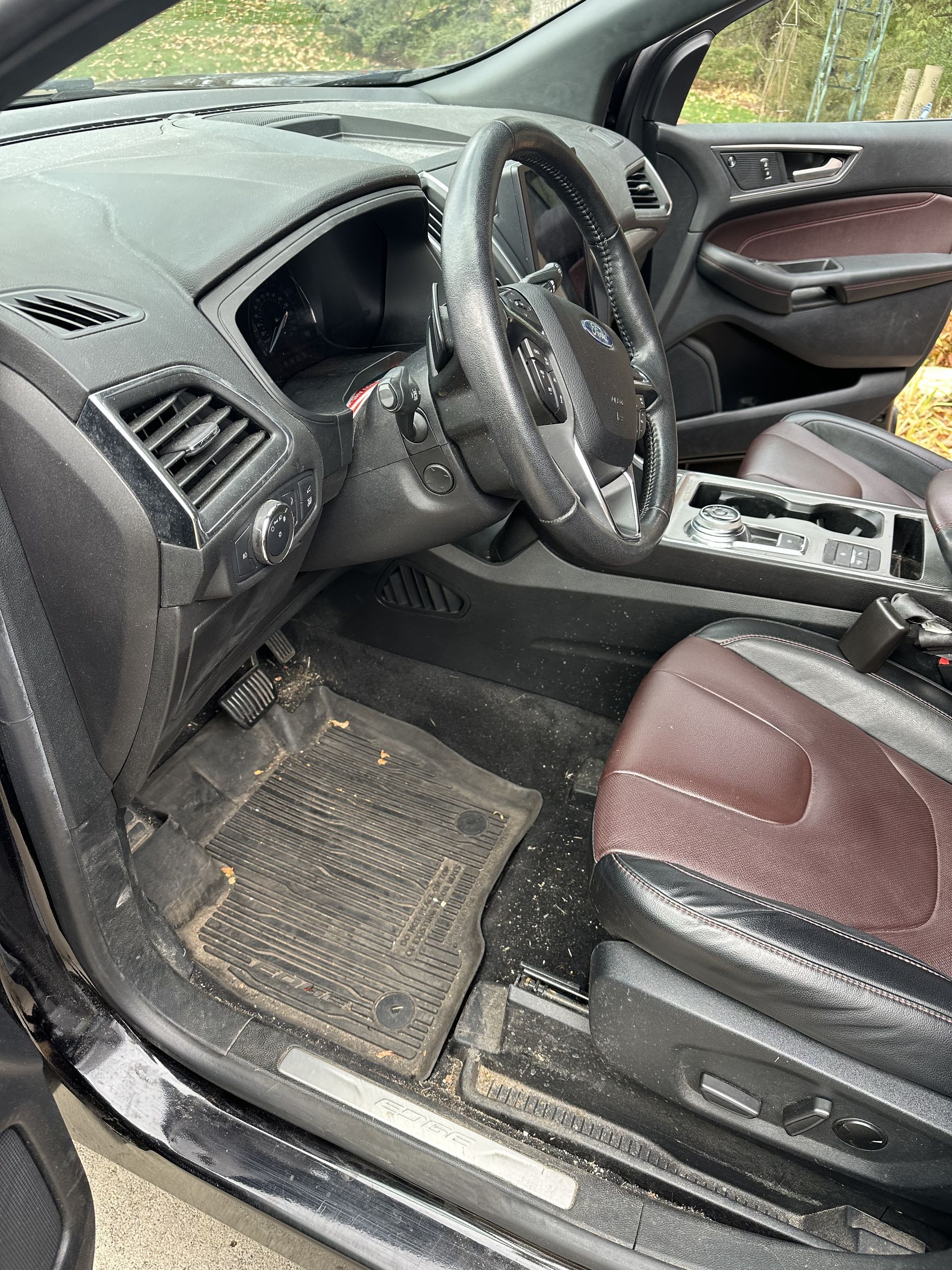 Interior of a car with a steering wheel, dashboard, black rubber floor mat, and maroon and black leather seats.