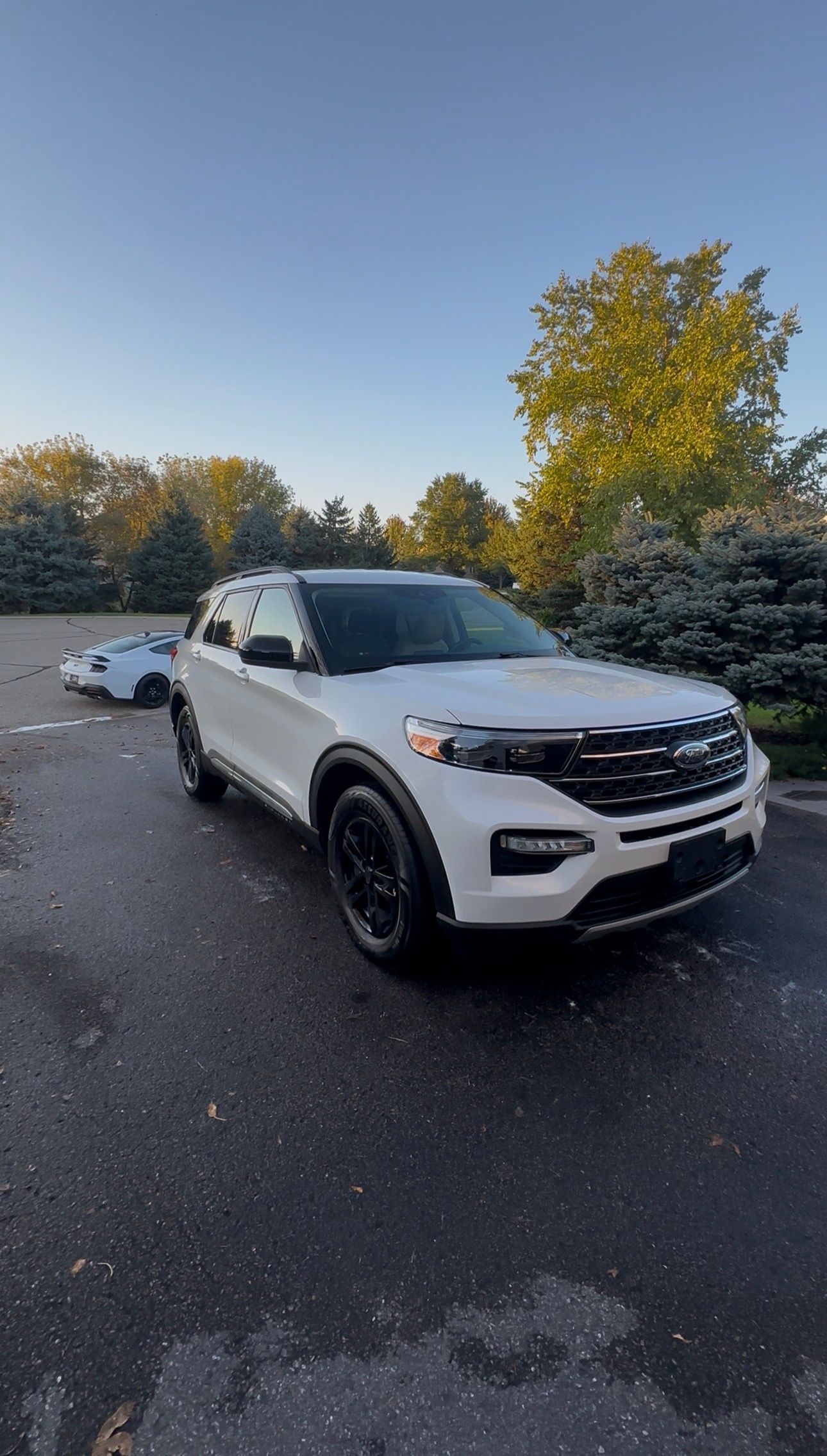 A white Ford Explorer SUV parked on a paved lot at sunset, with trees and a smaller car visible in the background.