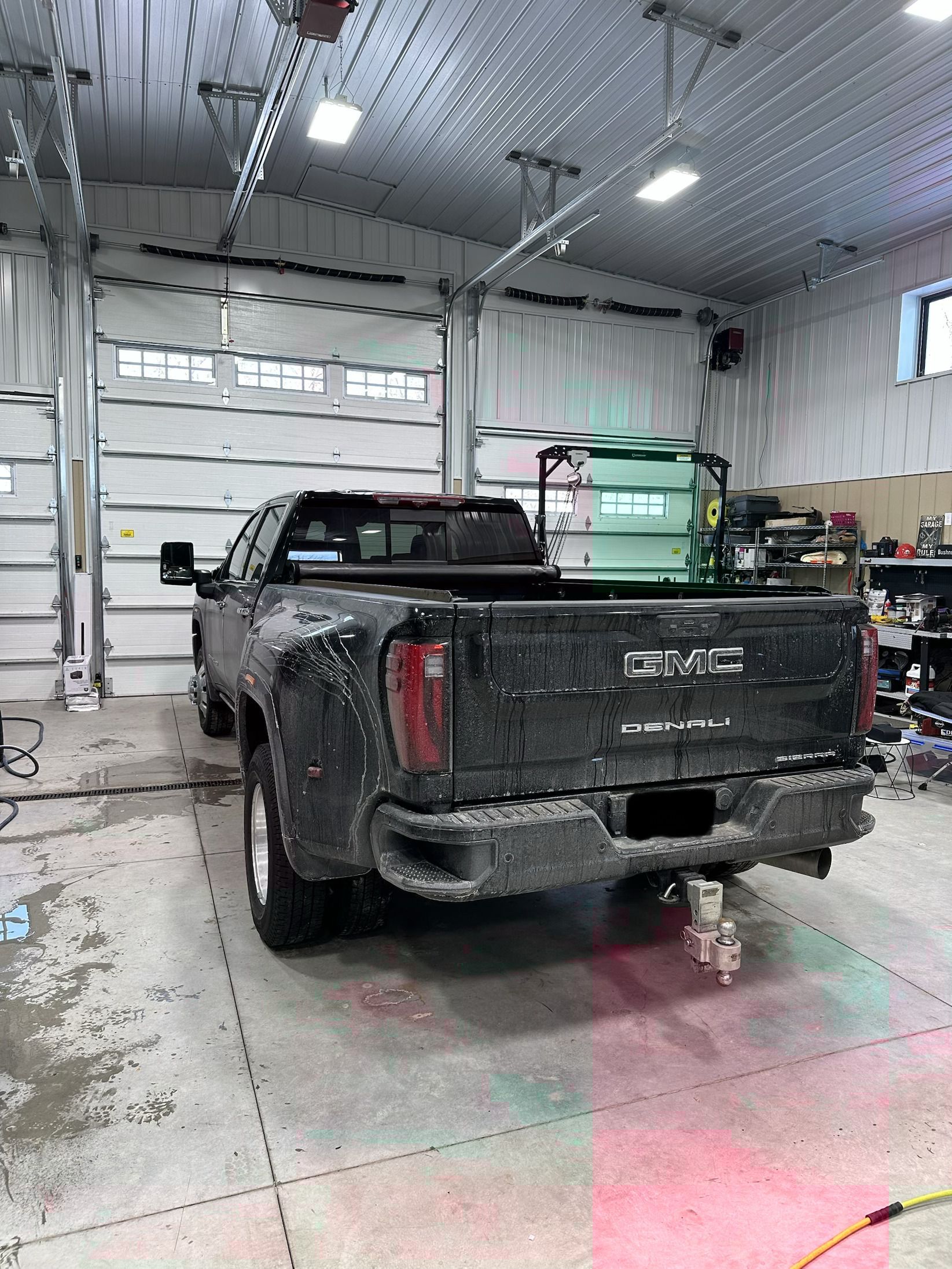 A mud-covered black GMC pickup truck parked in a large indoor workshop with white garage doors and bright lighting.