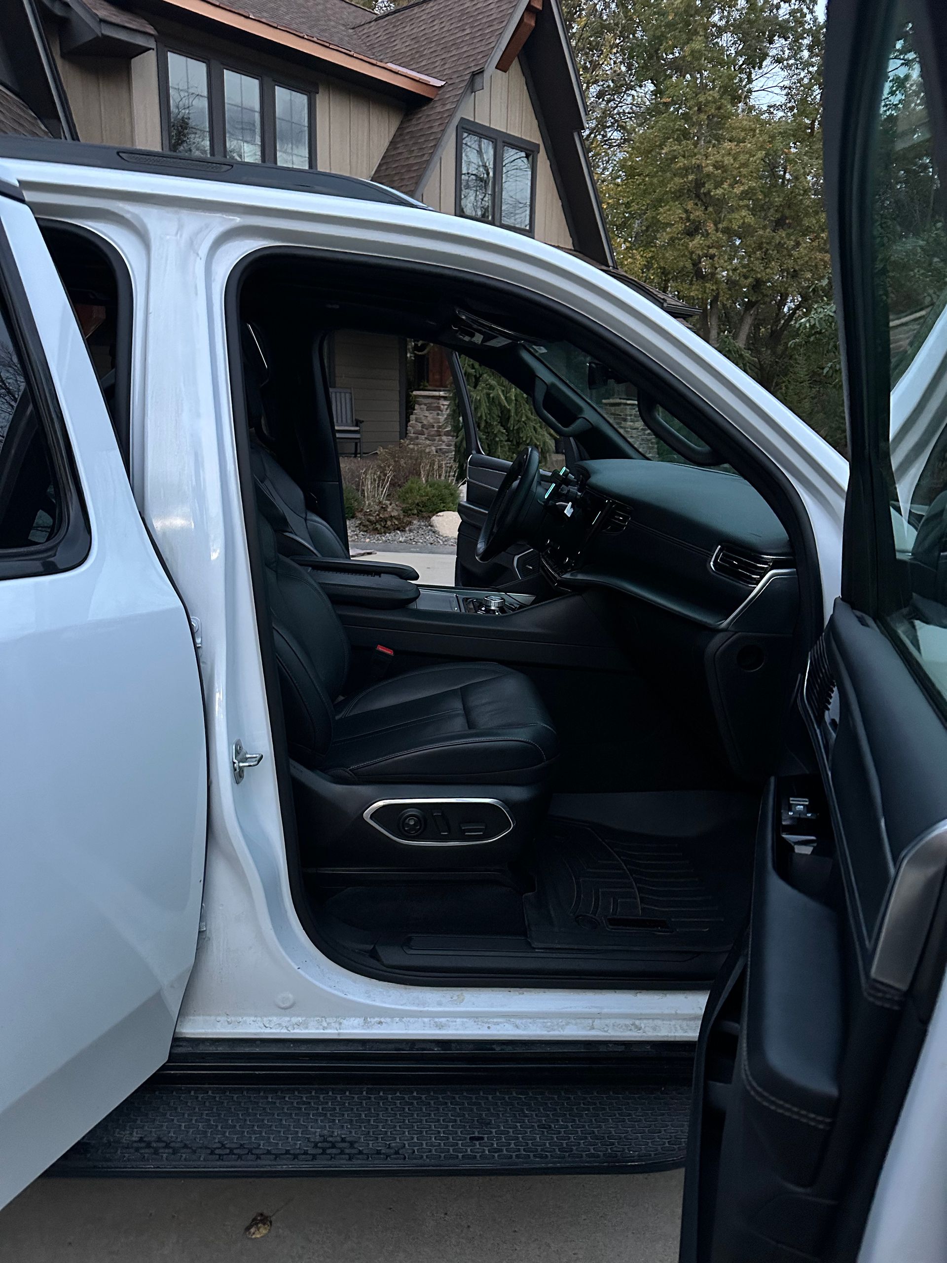 Open driver-side door of a white SUV, revealing black leather interior, seating, and dashboard, parked near a house.