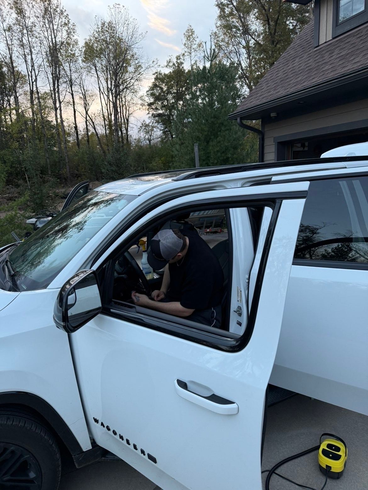 A person wearing a baseball cap works inside the front seat of a white SUV parked outside a home.
