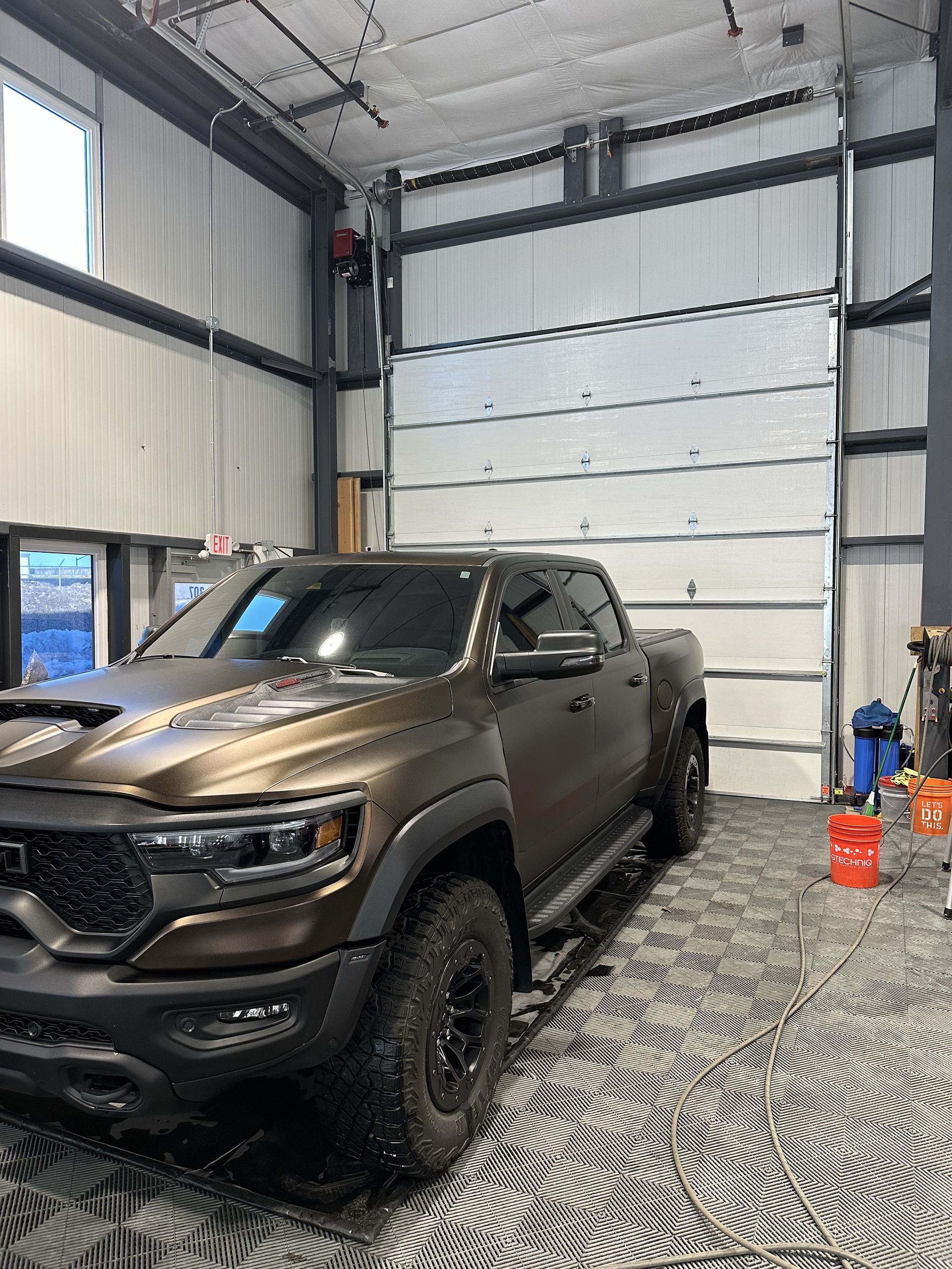 A matte bronze RAM TRX pickup truck parked inside a bright workshop with a patterned floor and large industrial door.