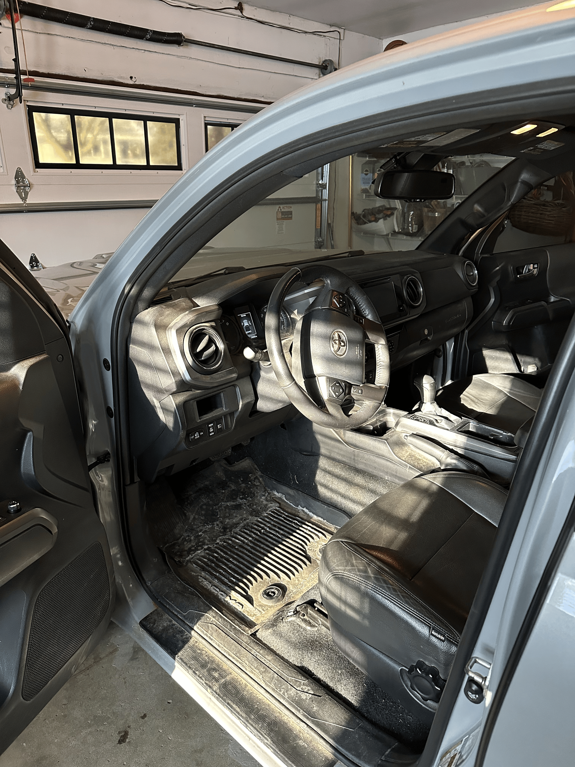 The interior of a vehicle, showing a steering wheel, dashboard, and floor mats covered in a thick layer of fine gray dust.