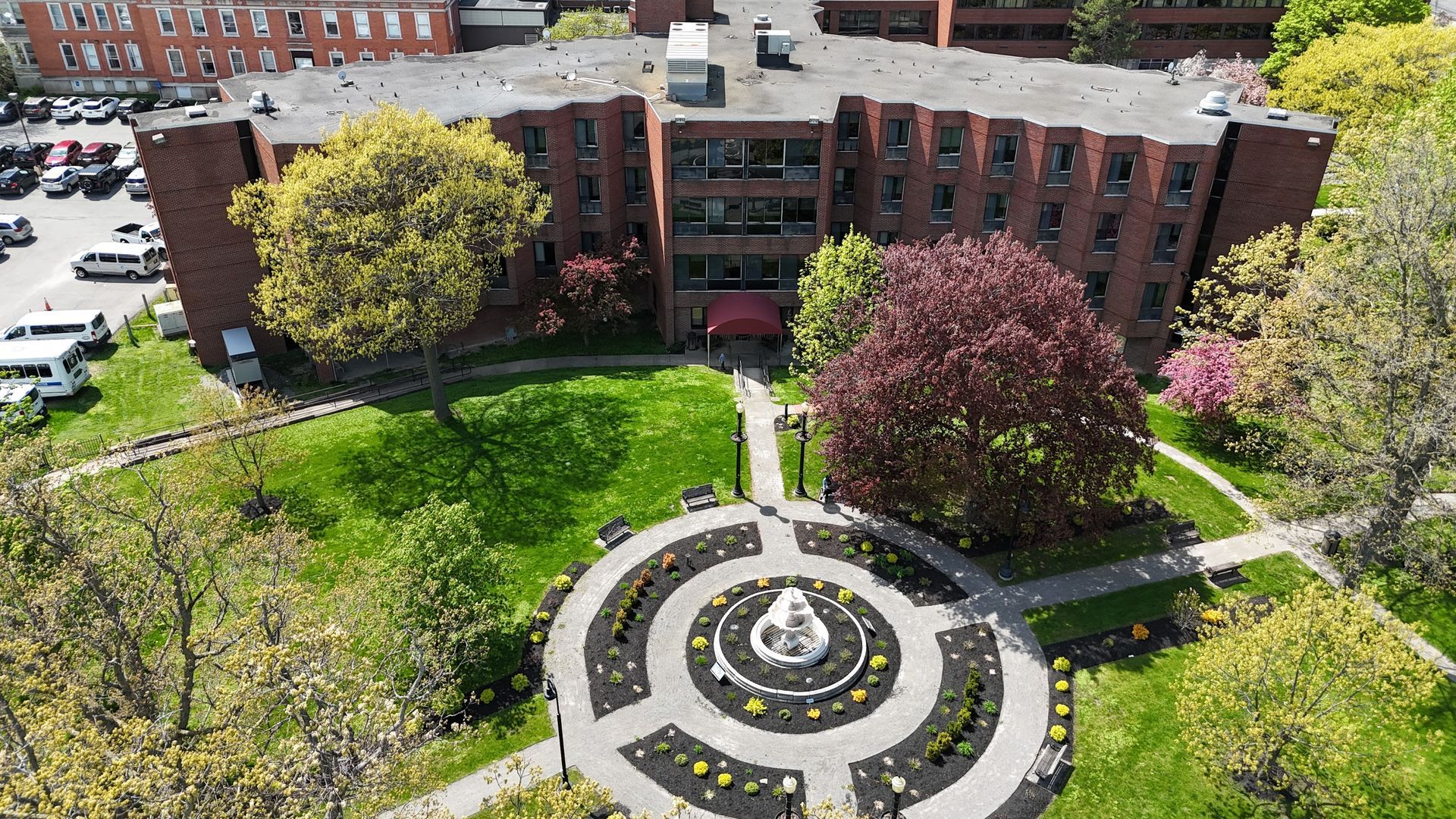 Aerial view of a circular garden fountain in a green courtyard beside a brick building and trees.