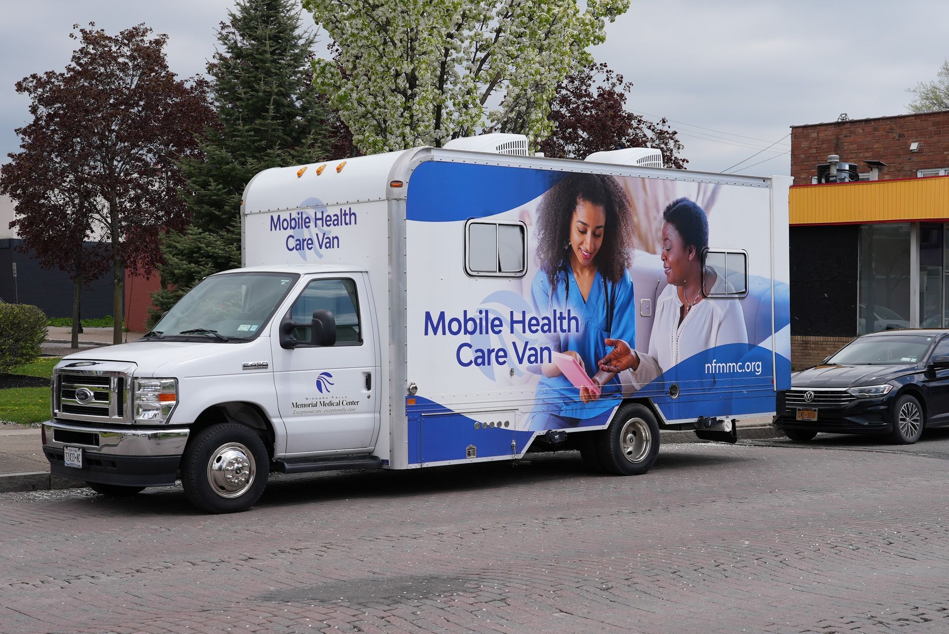 Mobile health care van parked on a street, with blue-and-white medical graphics and a nurse assisting a patient