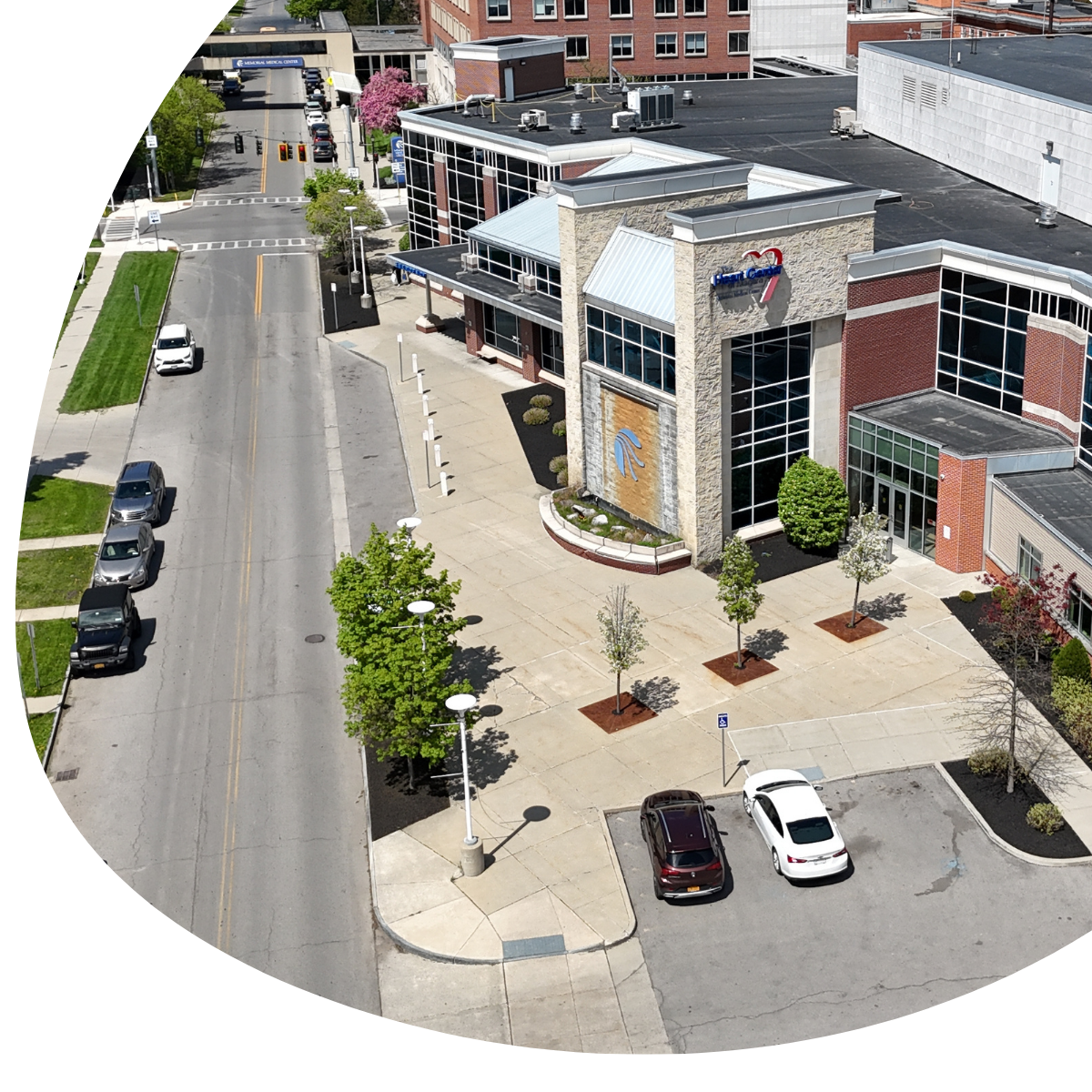 Aerial view of a modern building with glass walls beside a street and parking lot, with a few cars and trees.