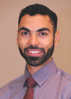 Smiling man in a lavender shirt and maroon tie against a beige background
