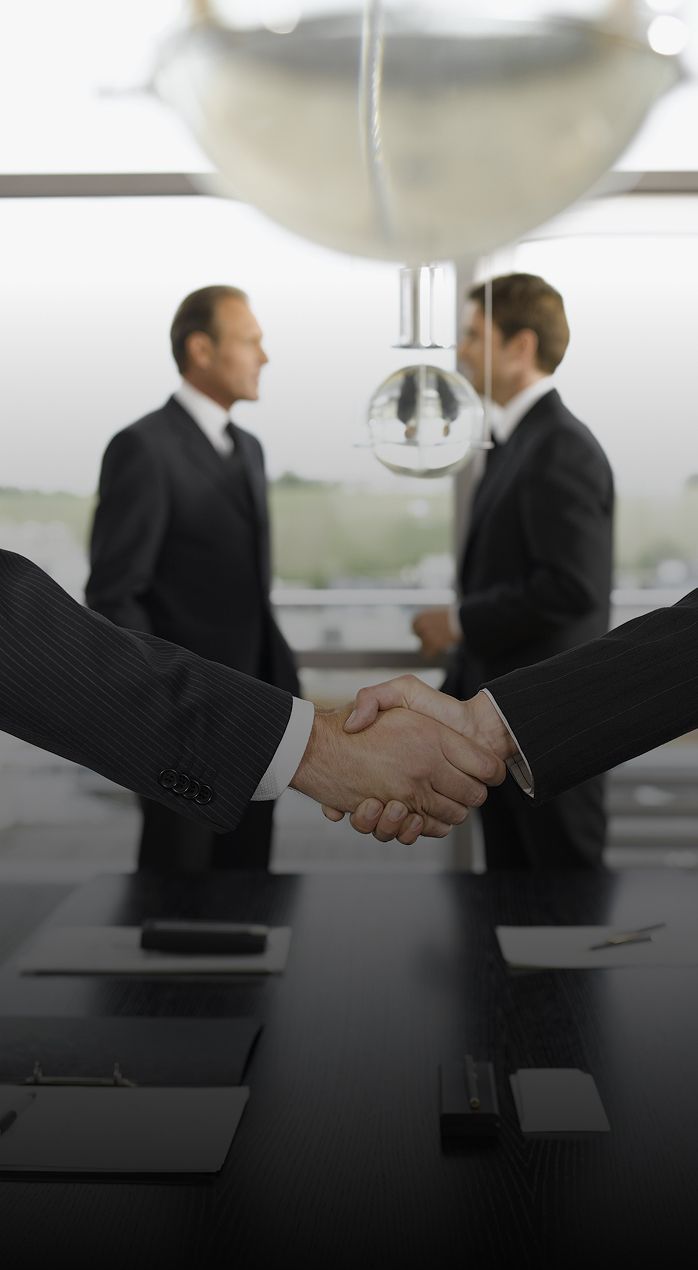 Two men shaking hands in an office, two others in suits stand in the background.