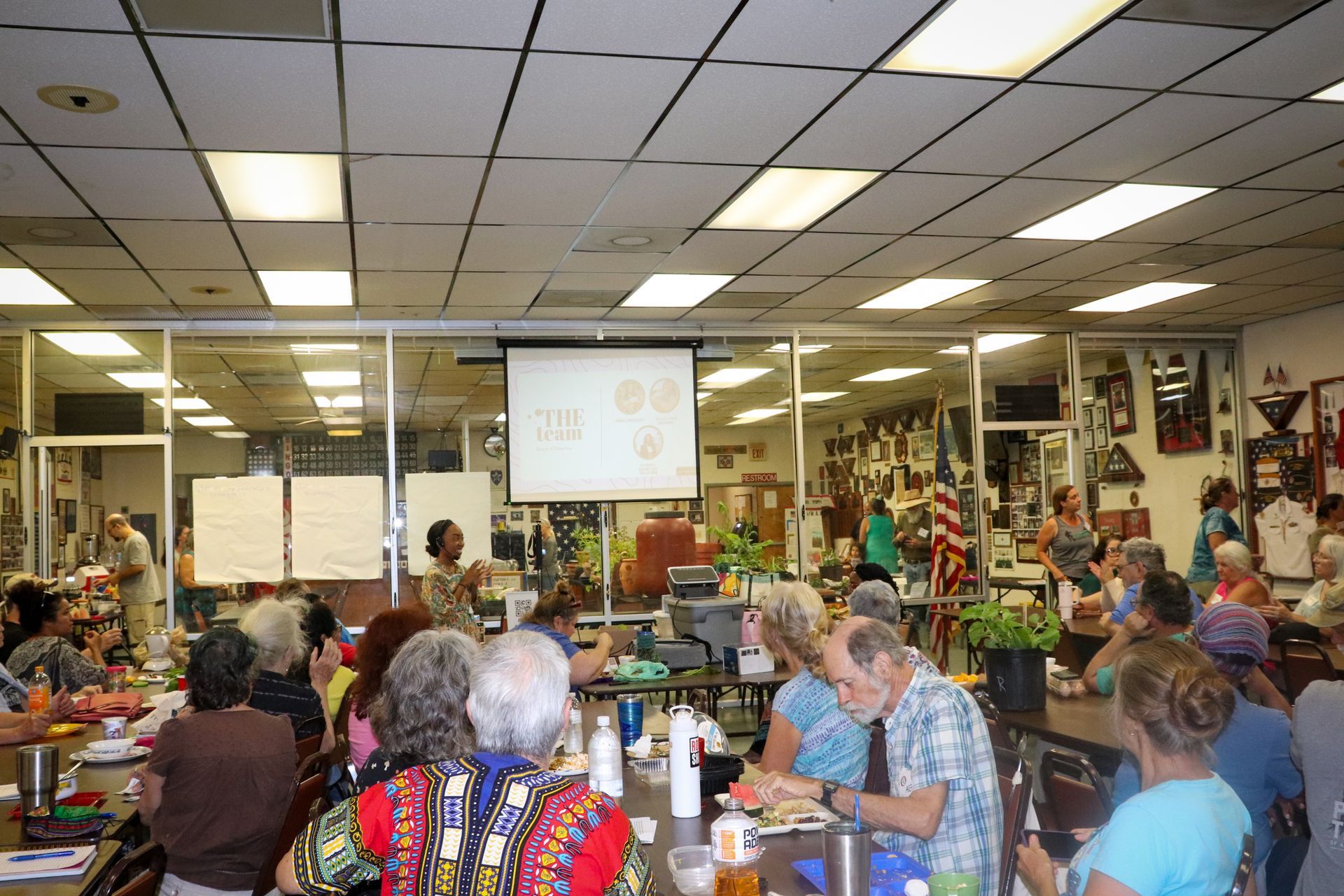 A large group of people are sitting at tables in a room with a projector screen.