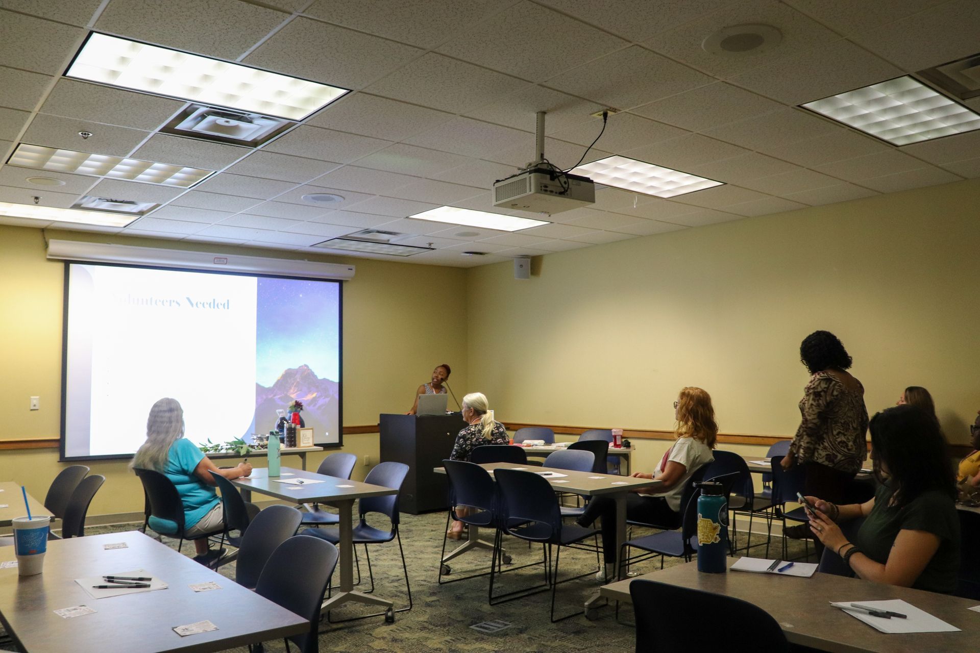 A group of people are sitting at tables in front of a projector screen.