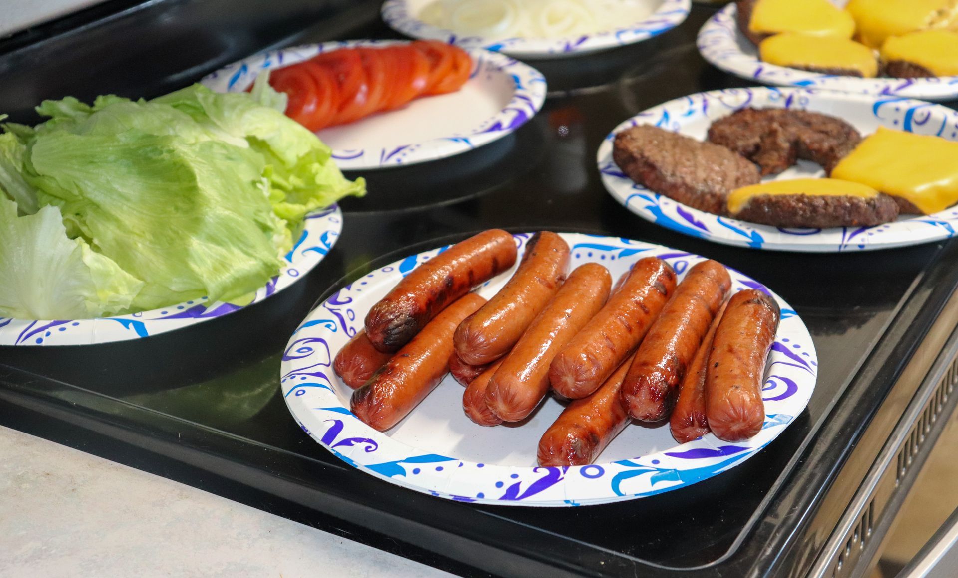 A bunch of plates of food on a stove including hot dogs and lettuce