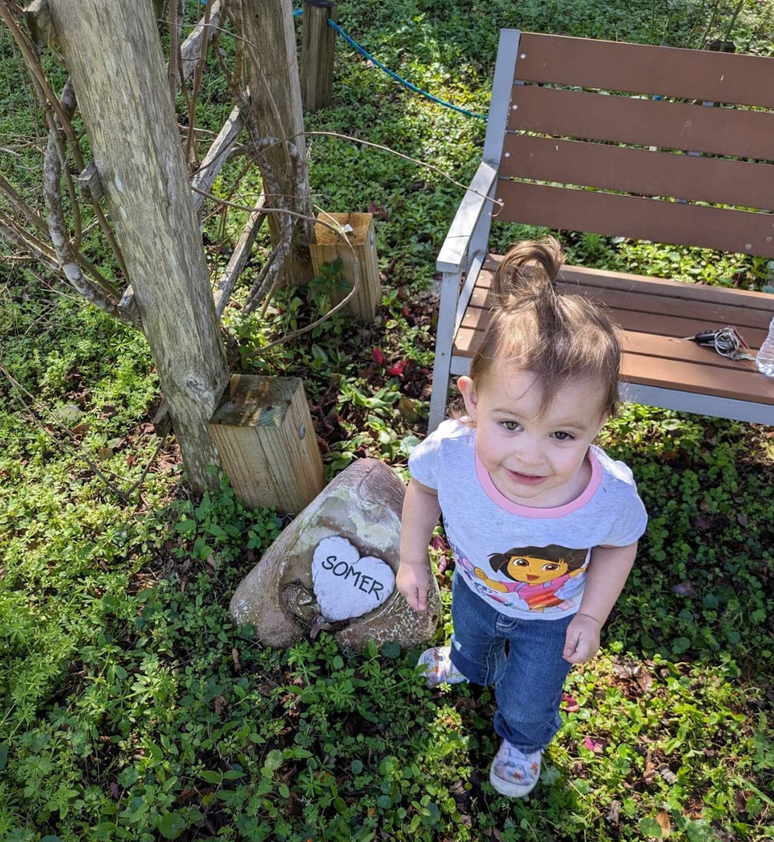 A little girl is standing in front of a wooden bench.