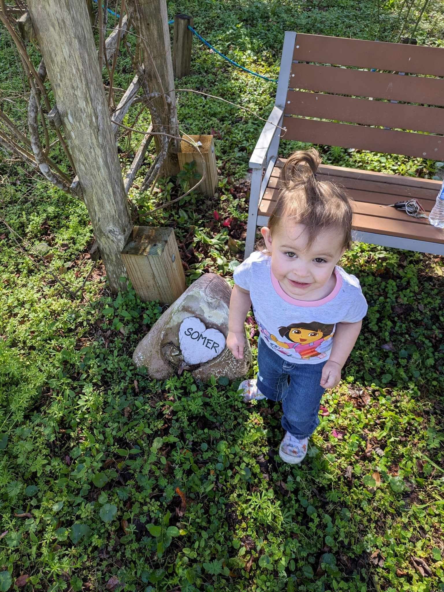 A little girl is standing next to a wooden bench in the grass.