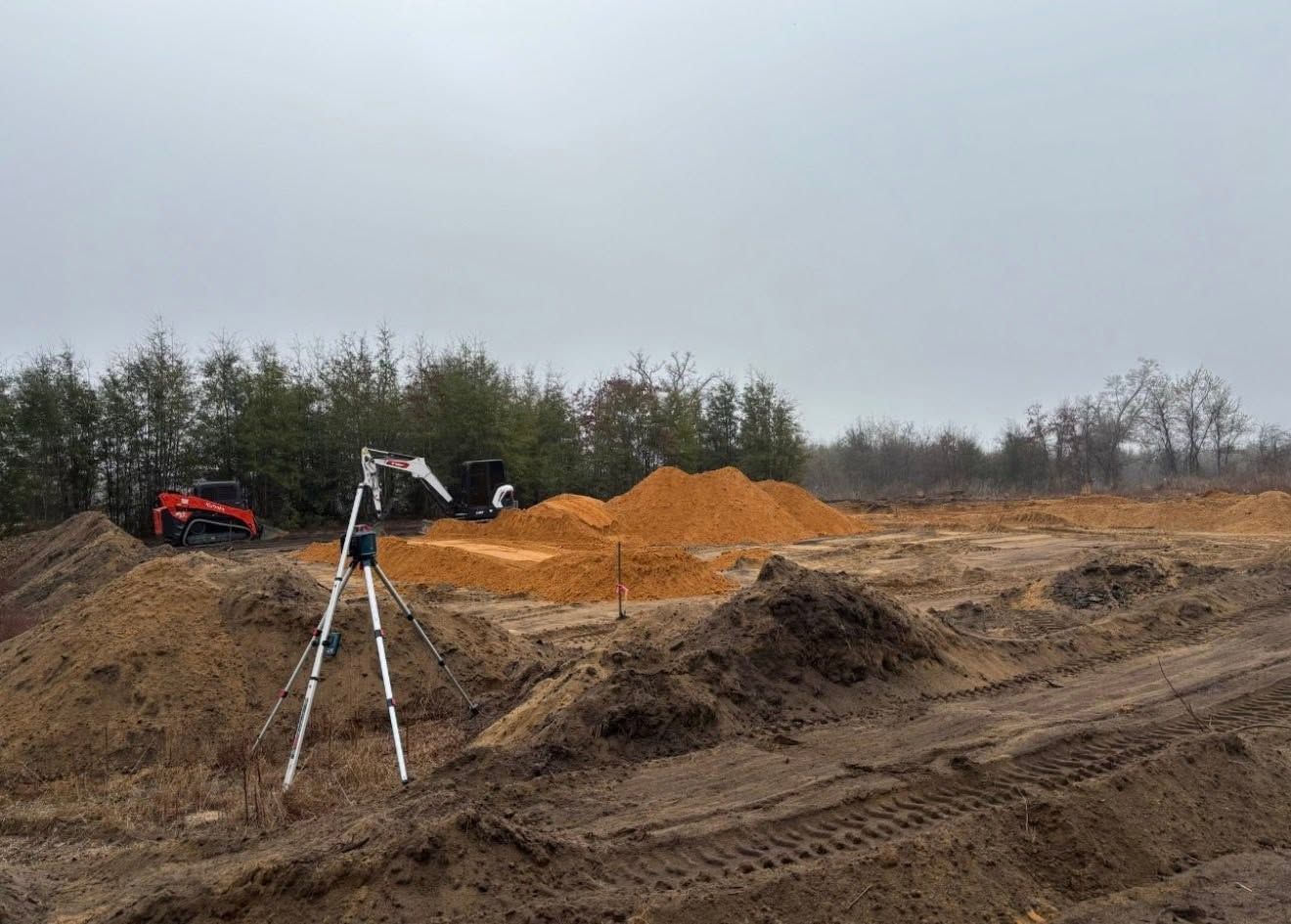 Construction site with orange dirt piles, a tripod survey marker, and a red excavator under cloudy sky