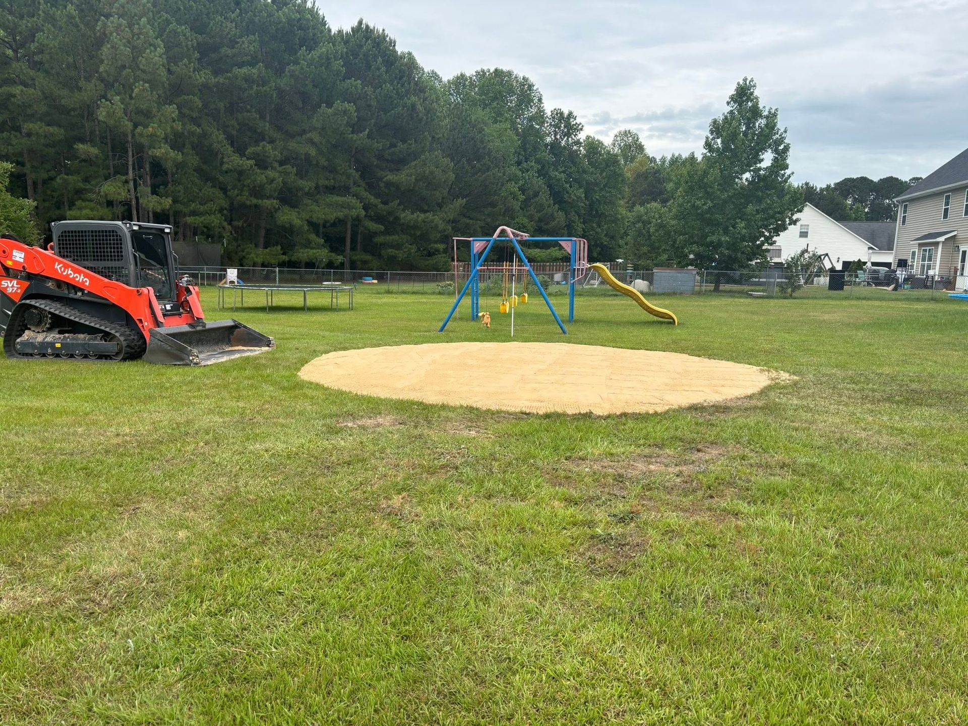 Small playground with a swing set and slide on a sandy area, next to an orange excavator in a grassy field.