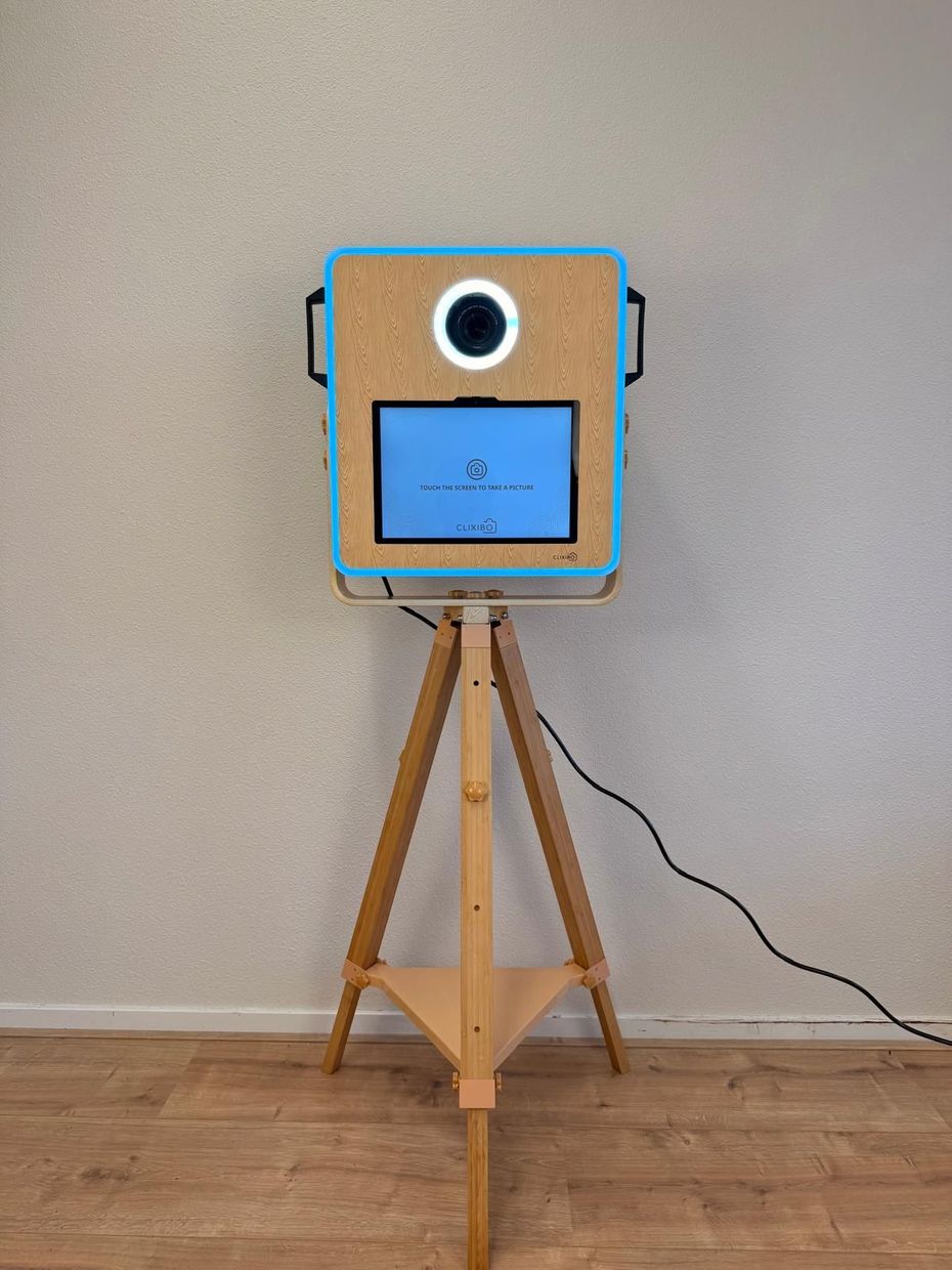 Een draagbare fotocabine met een tabletscherm en ringlamp, gemonteerd op een houten statief tegen een lichtwand. Photobooth