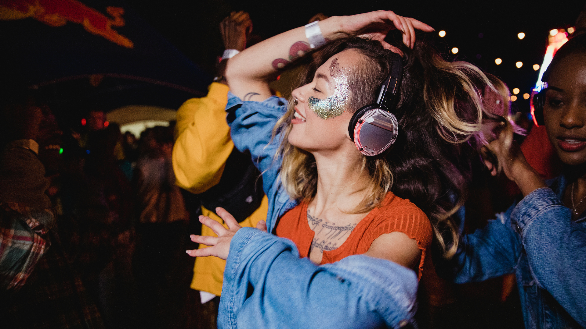 A woman wearing headphones and glitter on her face is dancing at a party.