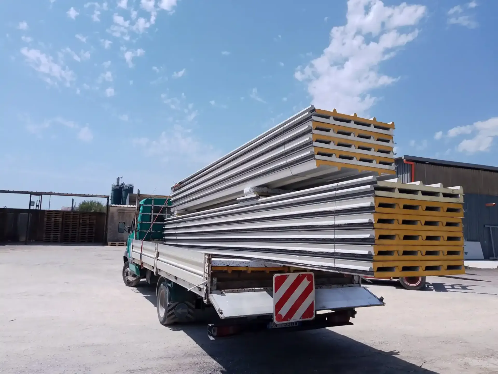 Un camion carico di pannelli metallici isolati, parcheggiato all'esterno di un edificio in una giornata di sole.