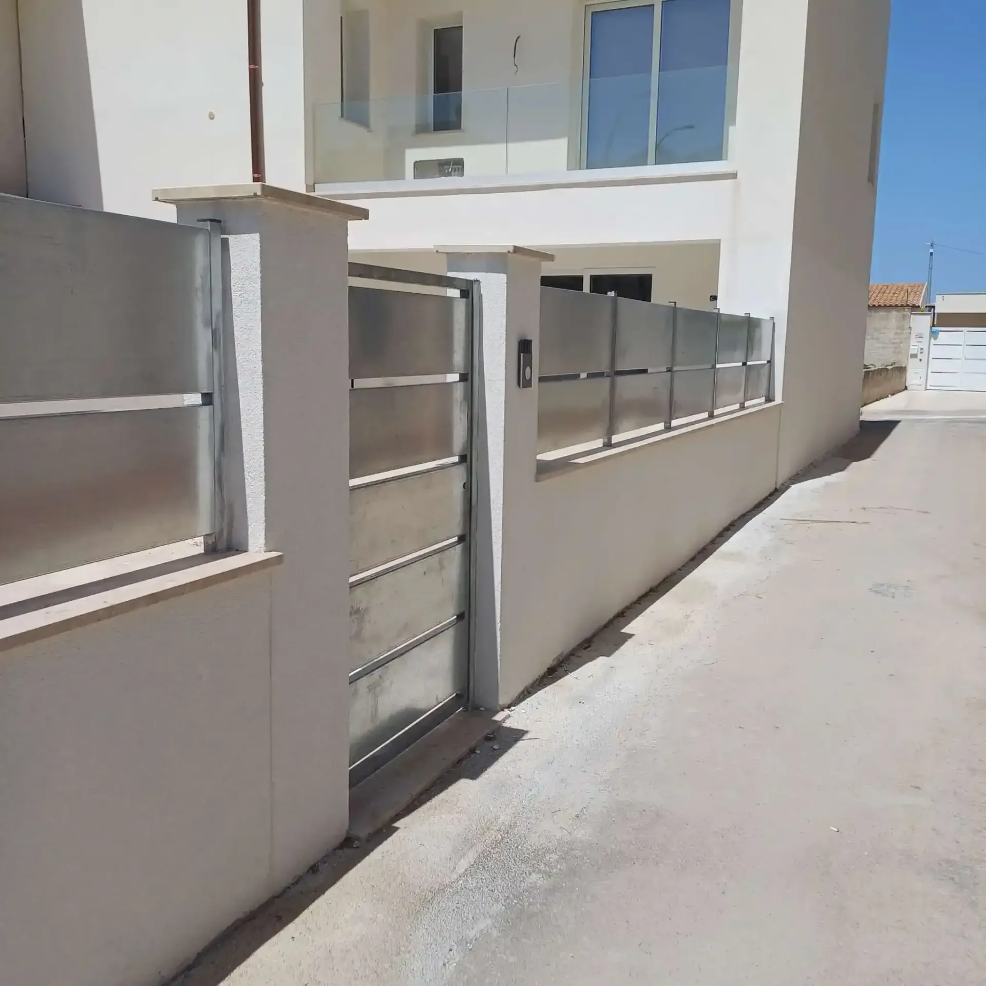 Una casa moderna con cancello in metallo e recinzione in vetro smerigliato. Pareti bianche, giornata di sole.