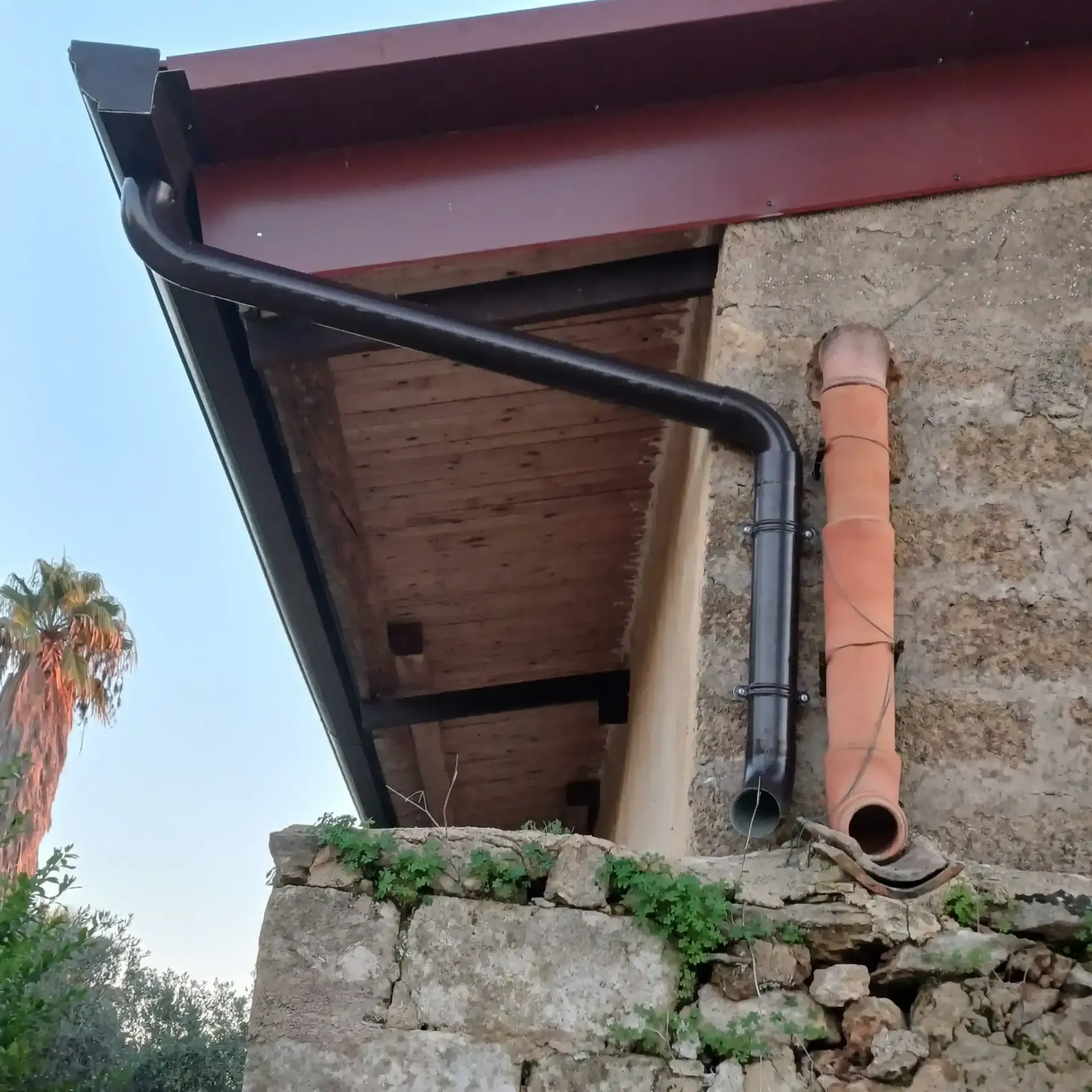 Sistema di grondaie nere su un edificio in pietra con tetto rosso-marrone e tubo in terracotta.