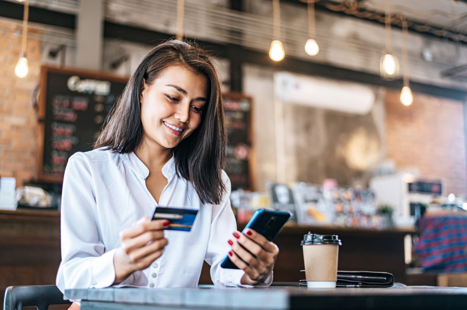 A woman is sitting at a table holding a credit card and a cell phone.