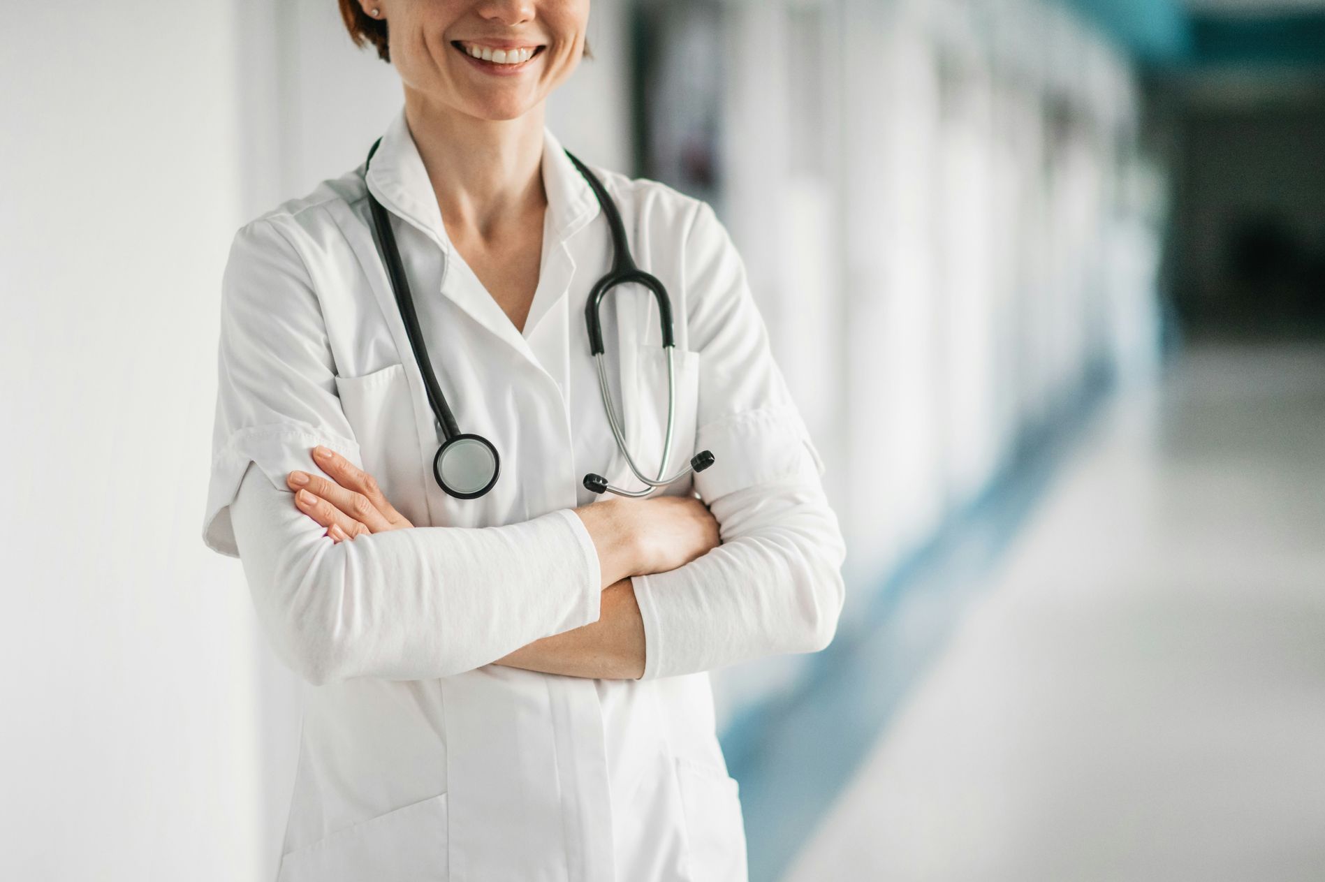 A female doctor with a stethoscope around her neck is standing in a hospital hallway with her arms crossed.