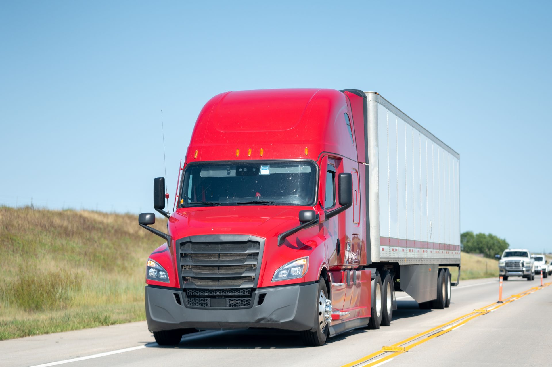 A red semi truck is driving down a highway next to a field.