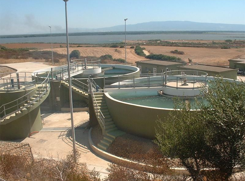 Impianto di trattamento delle acque con più serbatoi circolari, scale e uno specchio d'acqua sullo sfondo.