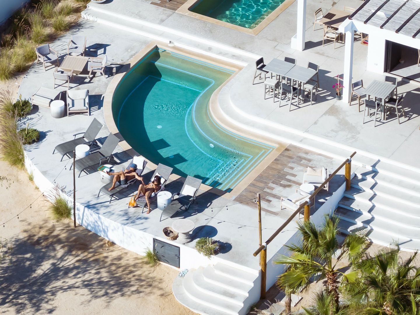 Aerial view of a white resort with multiple pools and lounge chairs; two people relax poolside.