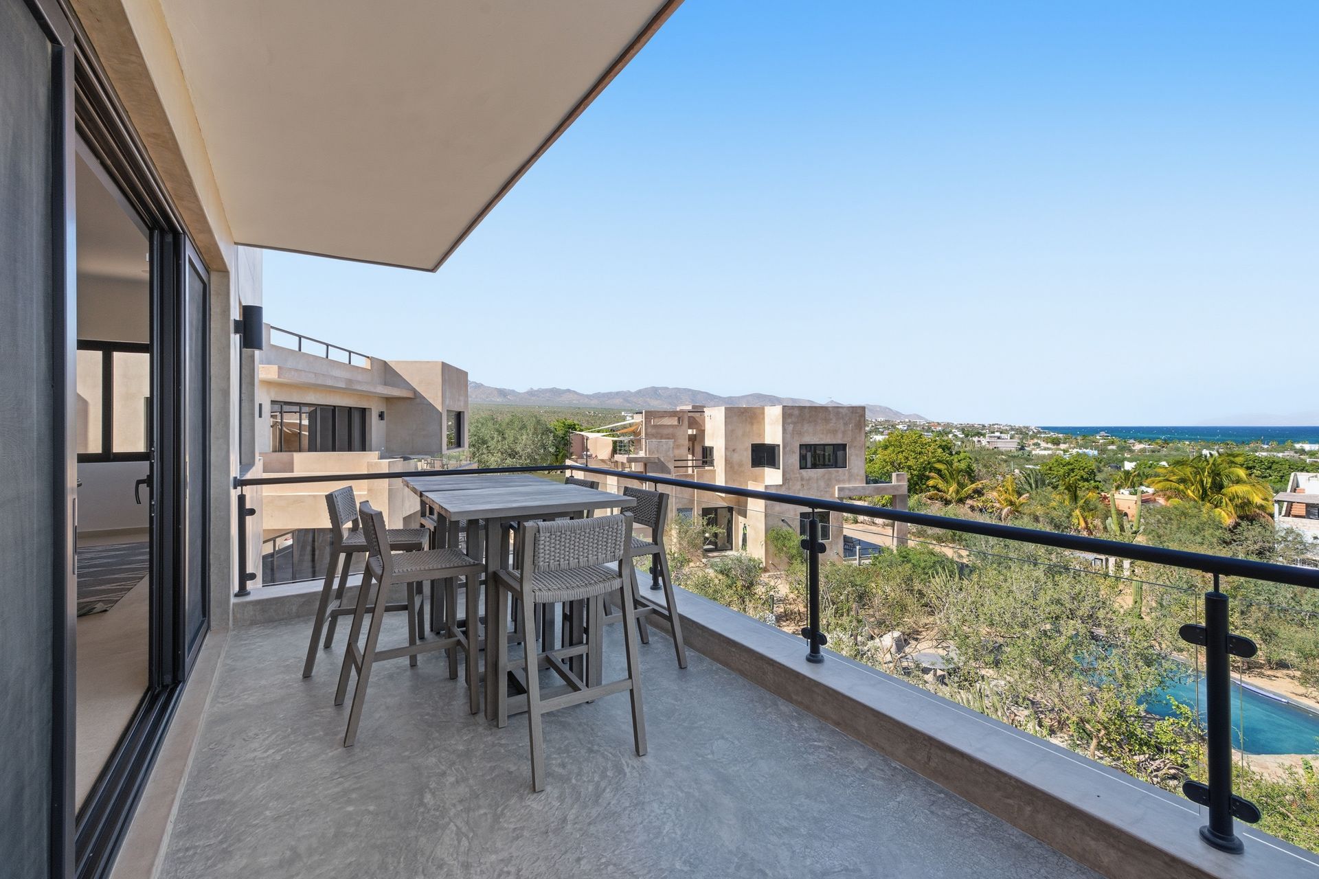 Balcony with table and chairs overlooking a landscape with buildings and a pool; bright blue sky.